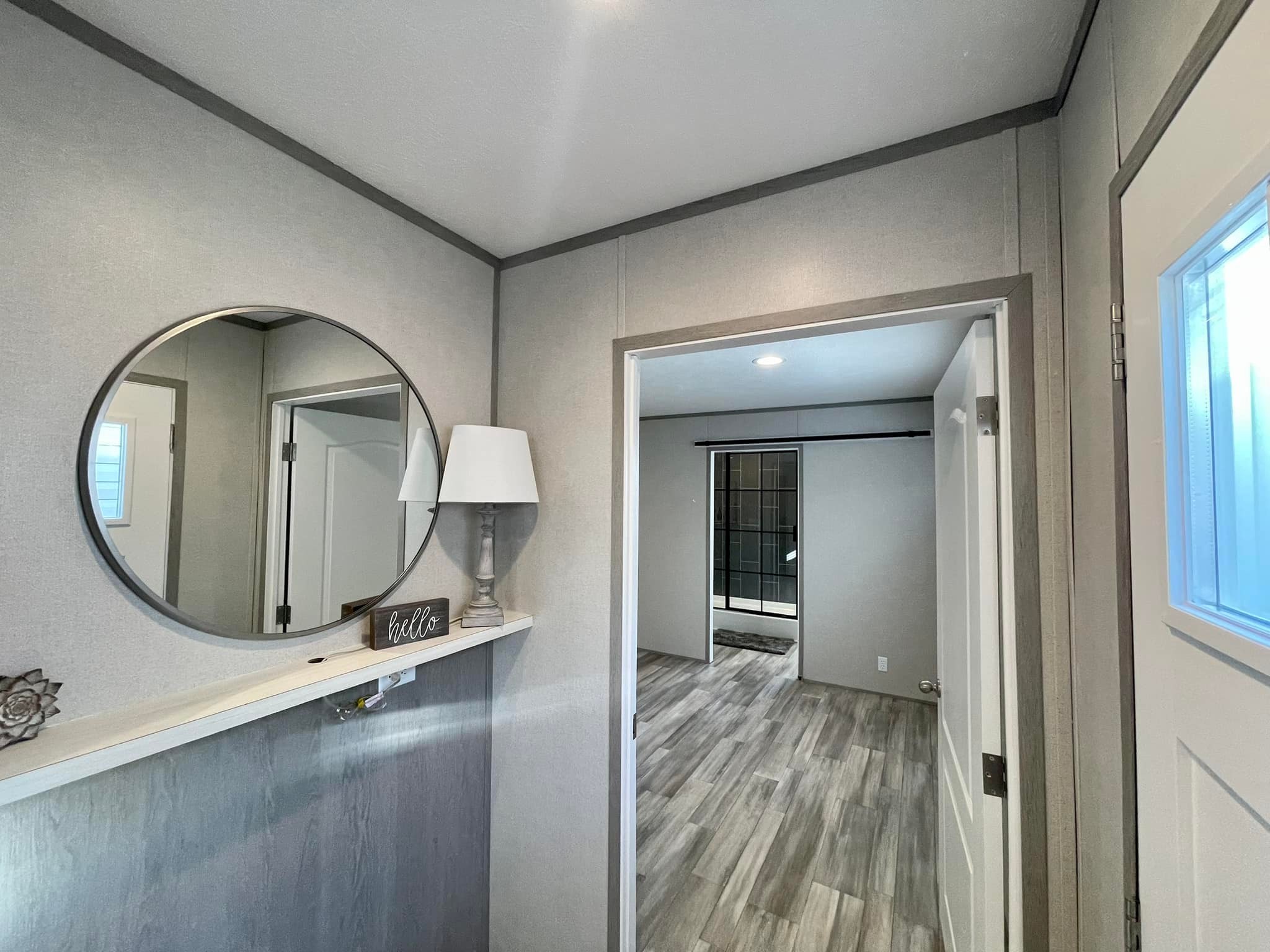 Small, modern entryway with gray walls, a round mirror, and a lamp on a shelf. An open door reveals a room with wood flooring and soft lighting.