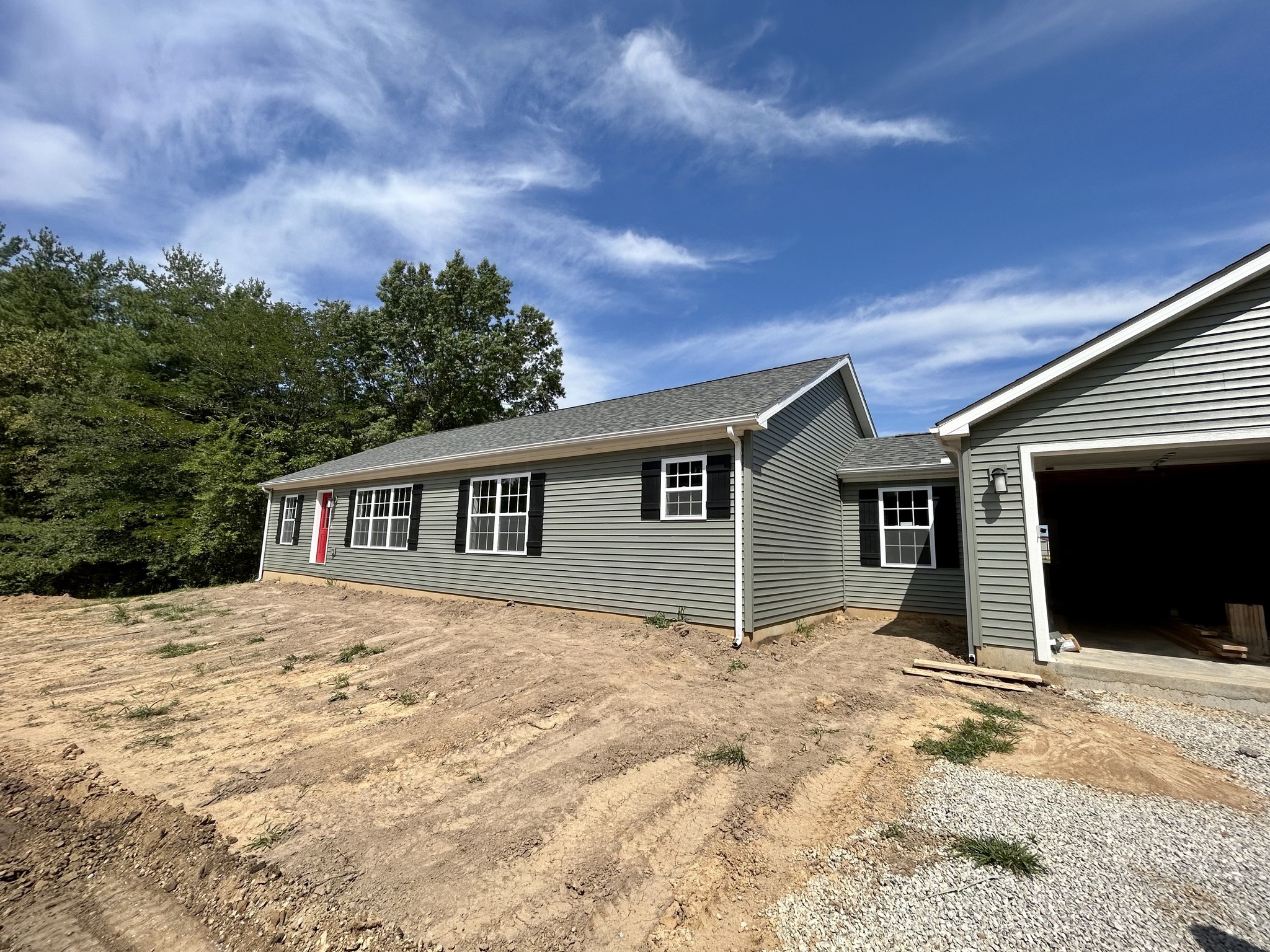 A single-story house with gray siding and black shutters, featuring a red door. A two-car garage is on the right, with a dirt yard in front. Clear blue sky above.