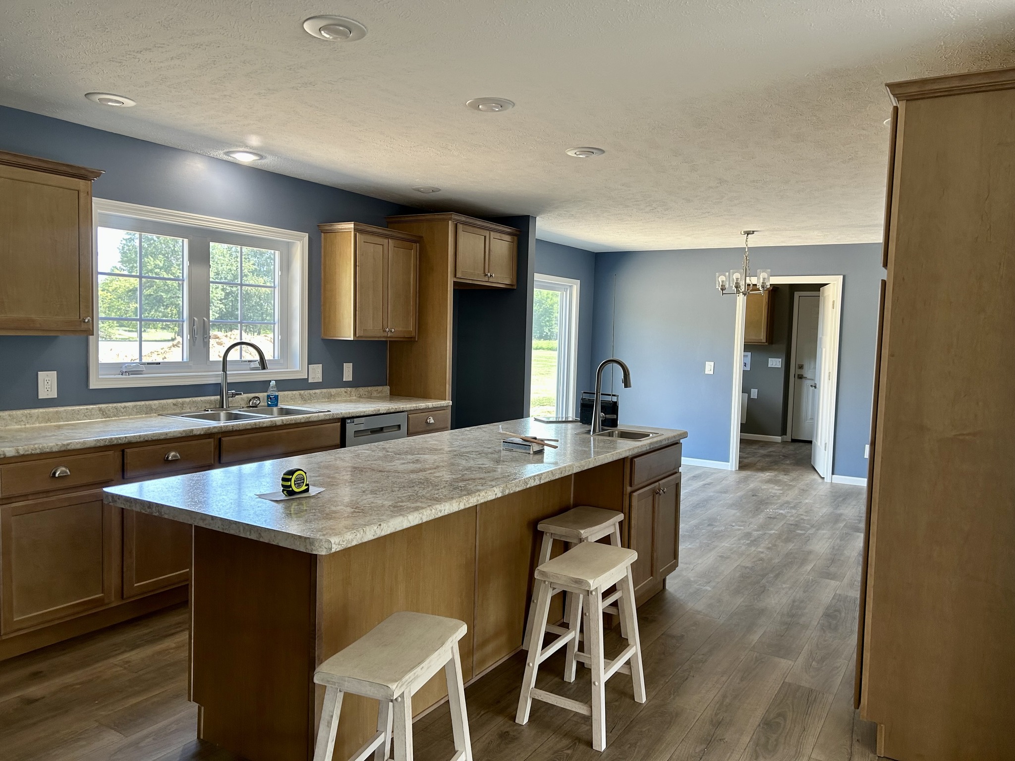 Bright kitchen with blue walls and brown wooden cabinets. A large island with a marble countertop and stools is in the center. Sunlight streams through a window.