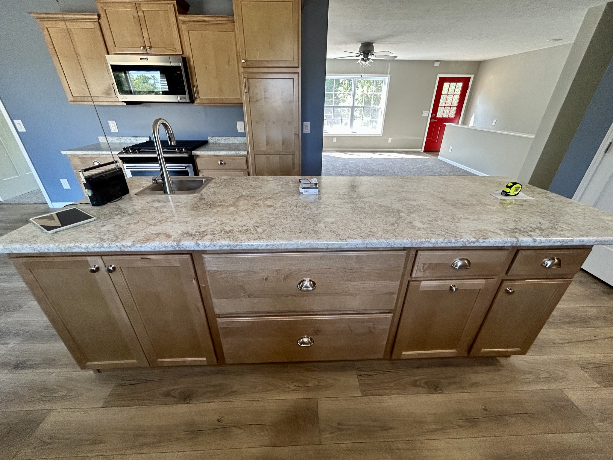 Modern kitchen with light wood cabinets and a large granite island. Sunlit, spacious with hardwood floors, stainless steel appliances, and a red door.