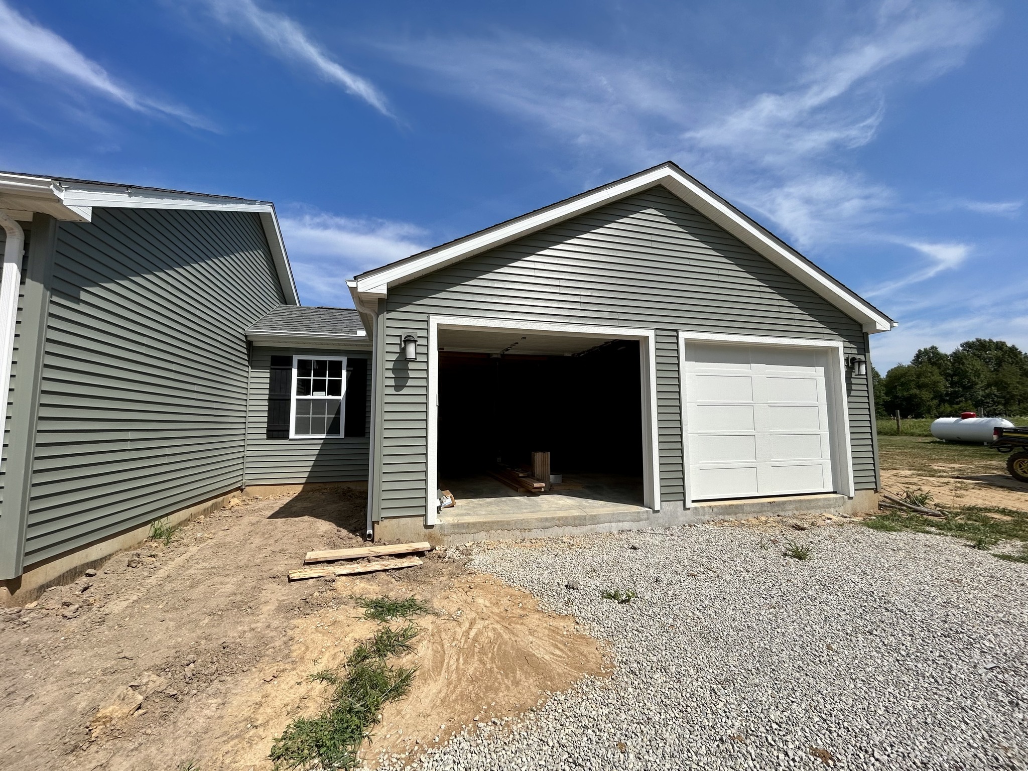 A newly constructed gray garage with white doors under a clear blue sky. Gravel driveway in front, surrounded by bare soil and wooden planks.