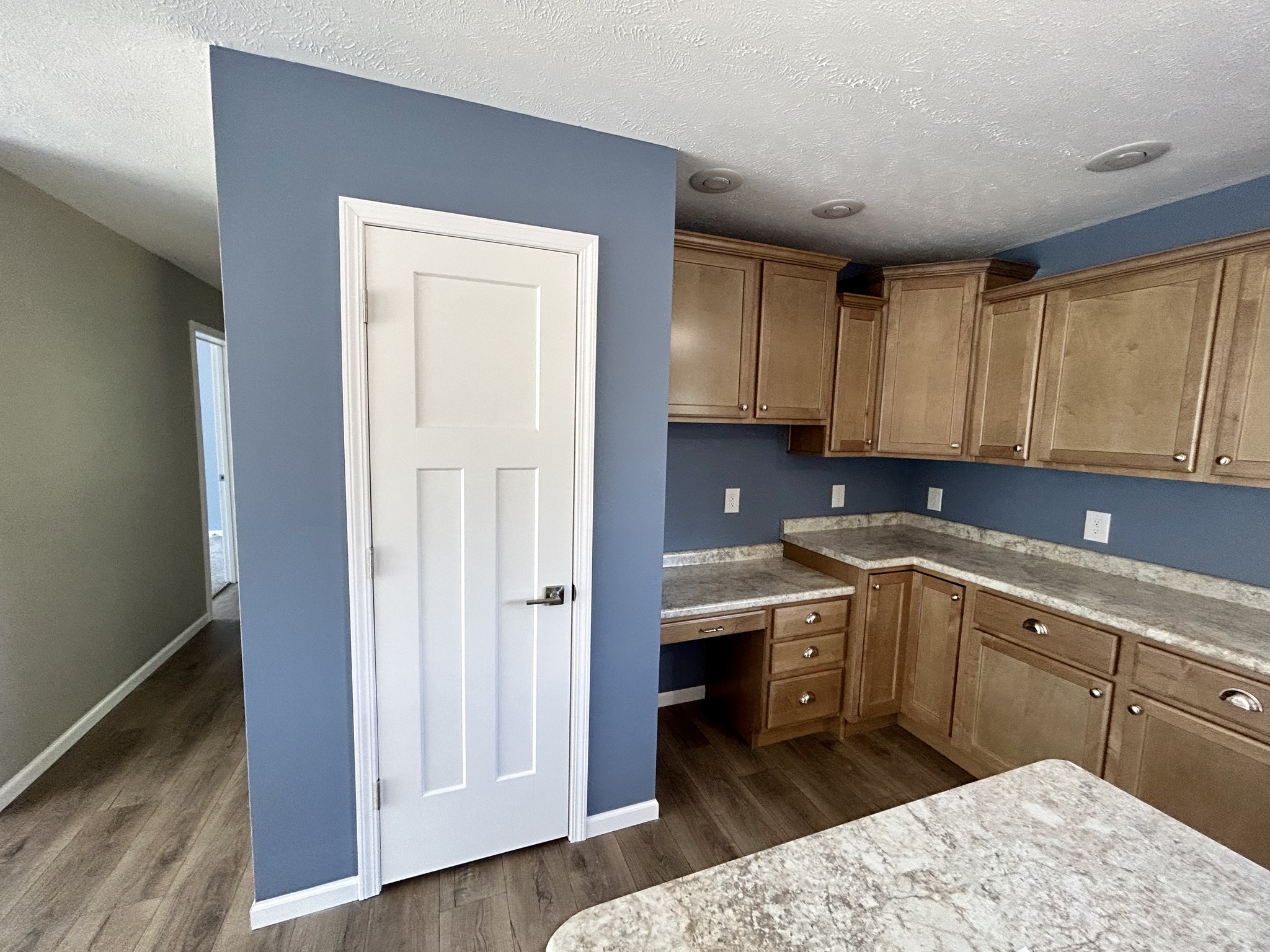 A modern kitchen with wooden cabinets, blue walls, and gray countertops. There's a white door on the left and hardwood flooring, creating a cozy feel.