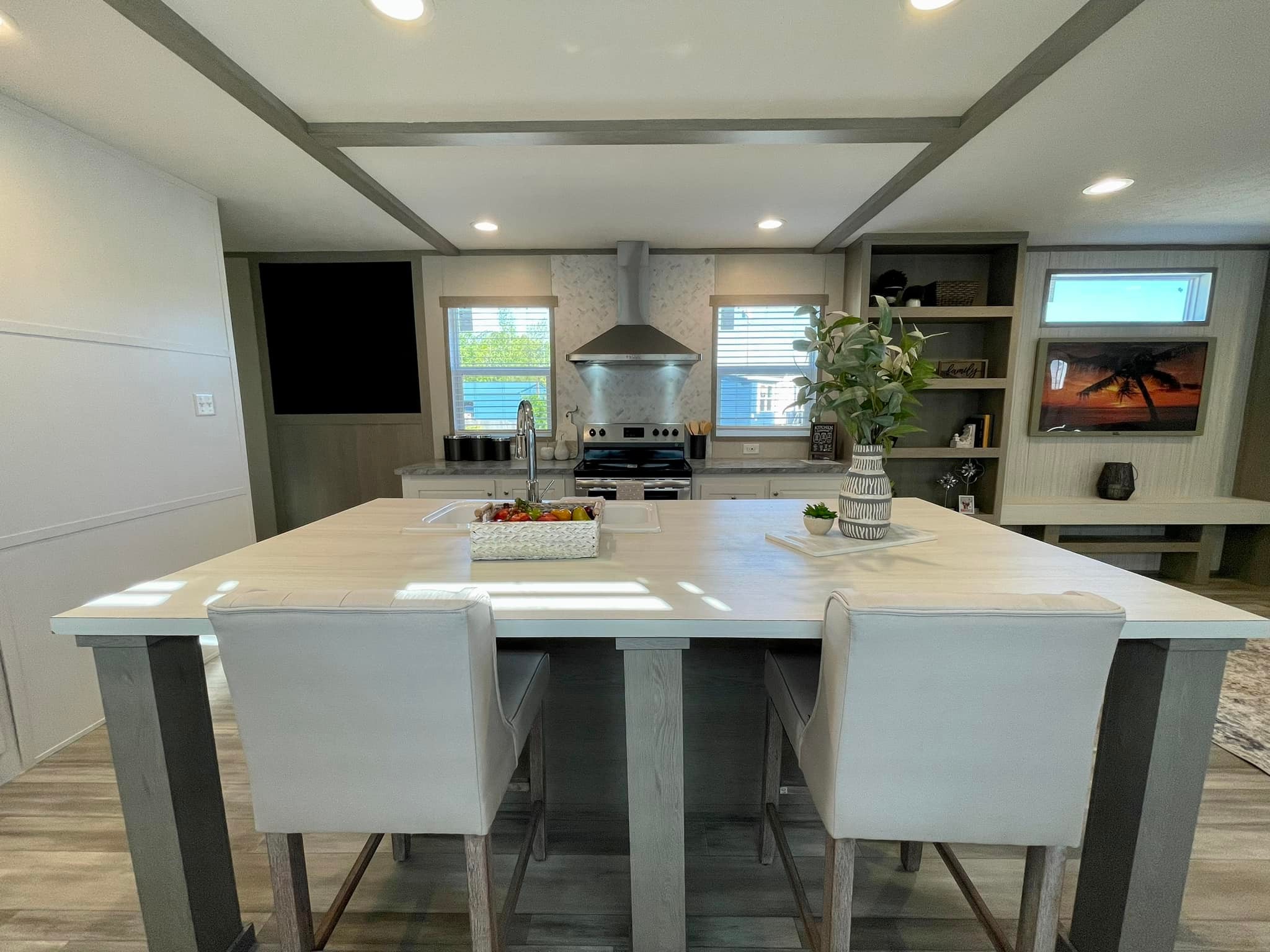 Modern kitchen with a large island featuring white countertops and two cushioned bar stools. Stainless steel stove, range hood, and vase with flowers add elegance. Bright and welcoming atmosphere.