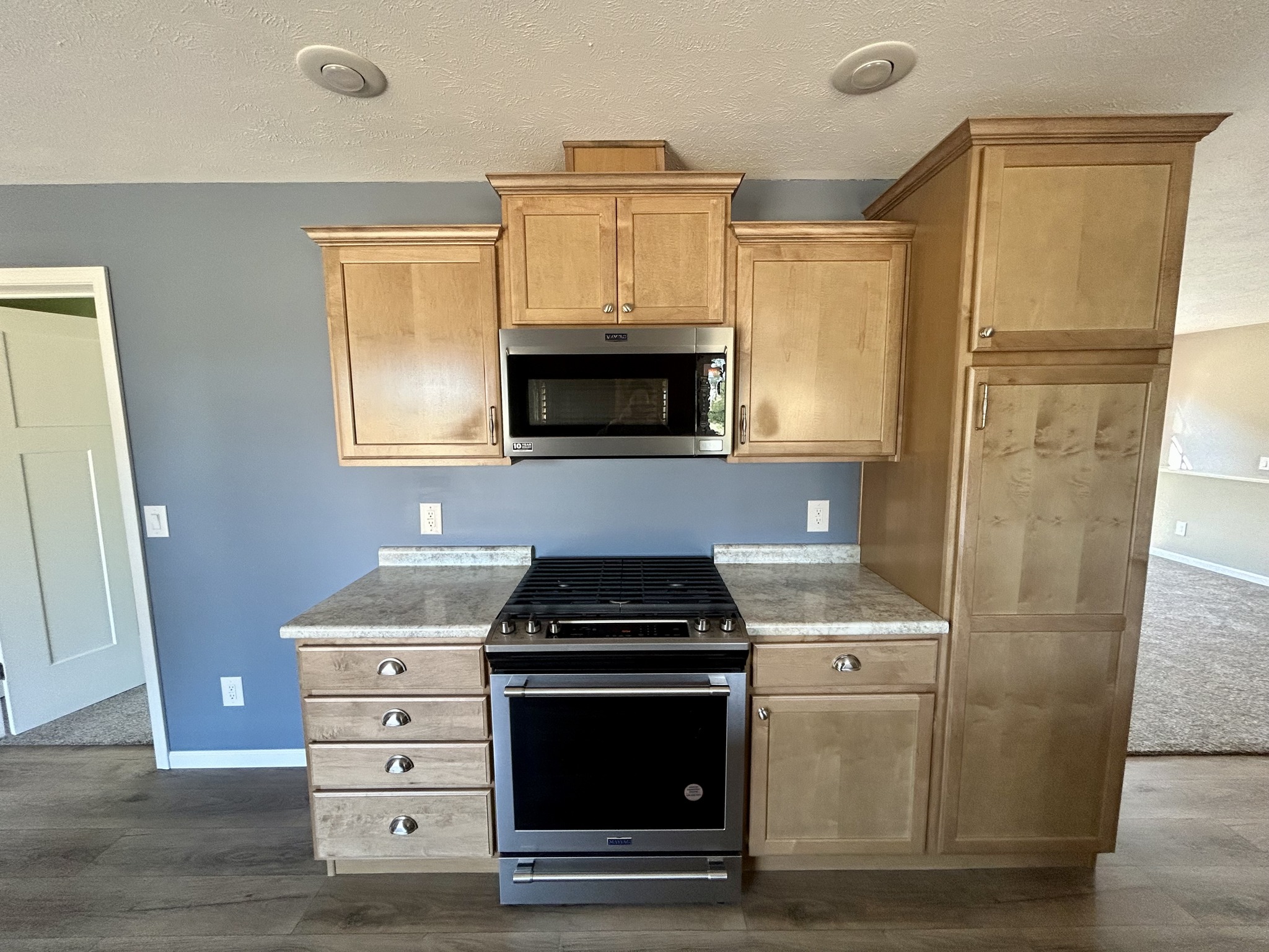Modern kitchen with light wood cabinets, stainless steel microwave, and a gas stove. Blue-grey wall adds contrast, creating a sleek look.