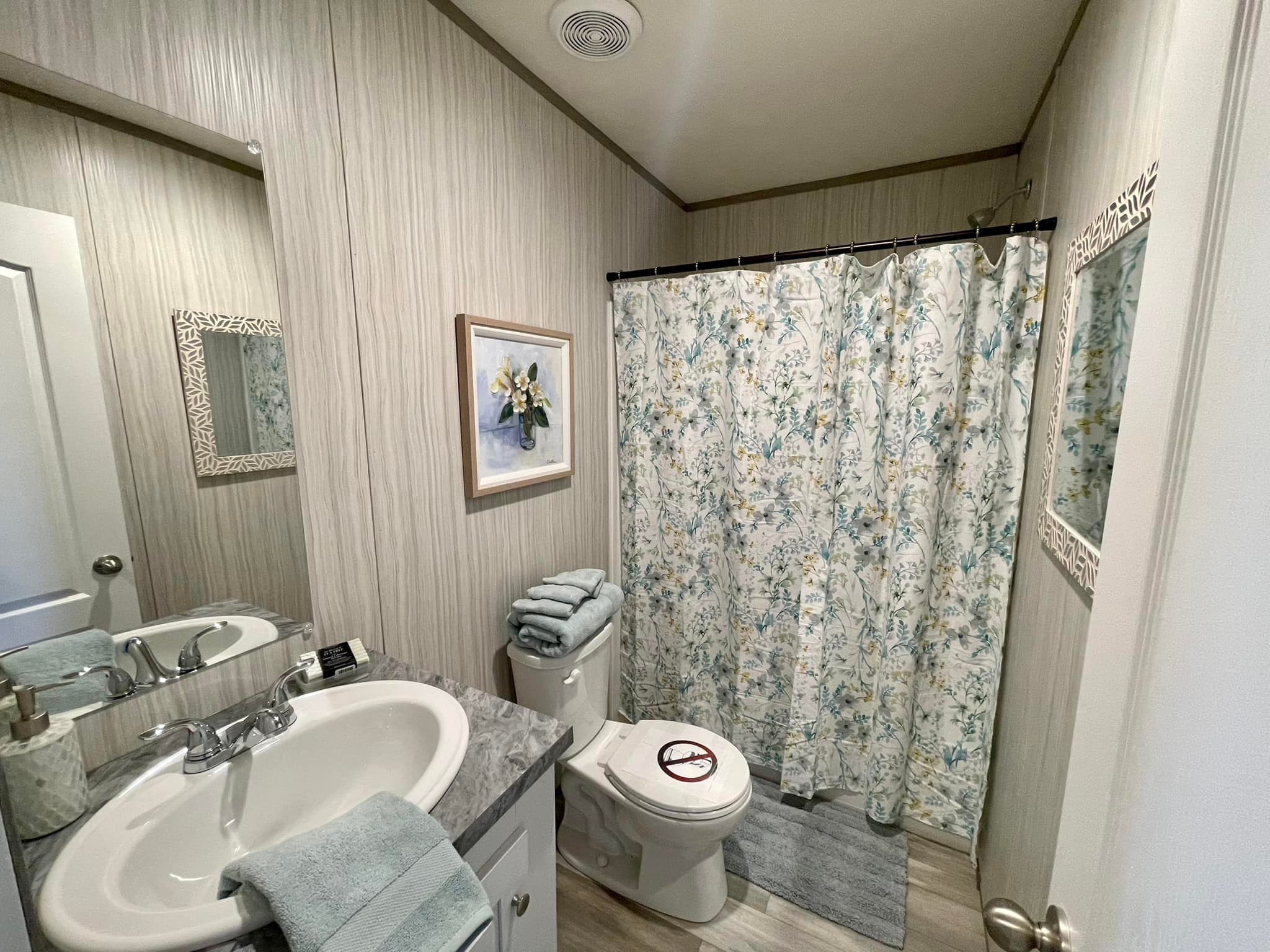 A bathroom with light wood-patterned walls features a sink with a silver faucet, a floral shower curtain, a toilet, and neatly folded towels. The decor includes a floral painting and a decorative mirror, conveying a serene, tidy atmosphere.