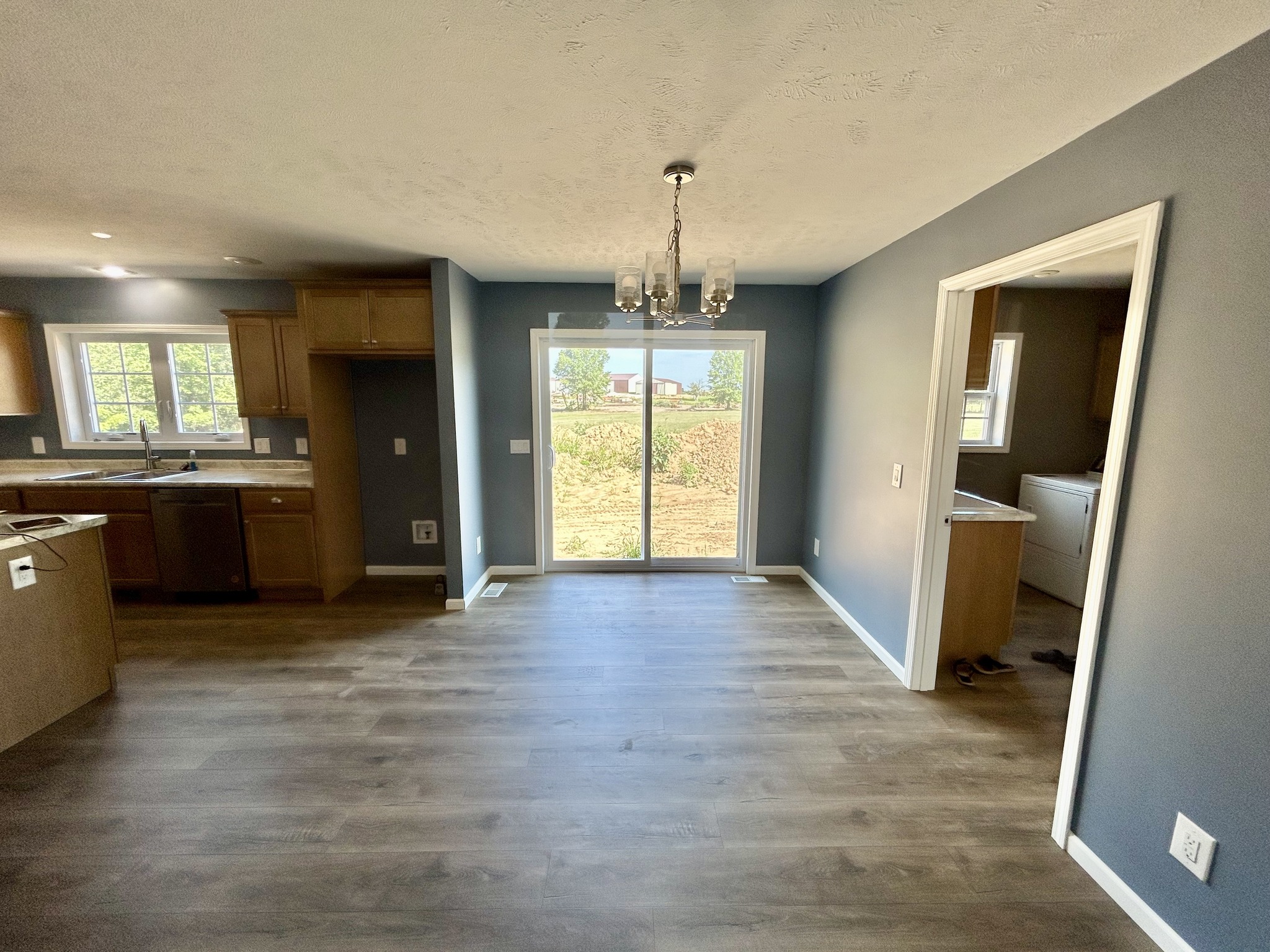 Spacious room with gray walls, wooden floor, and modern chandelier. Features a sliding glass door opening to a garden view, adjacent to a kitchen.