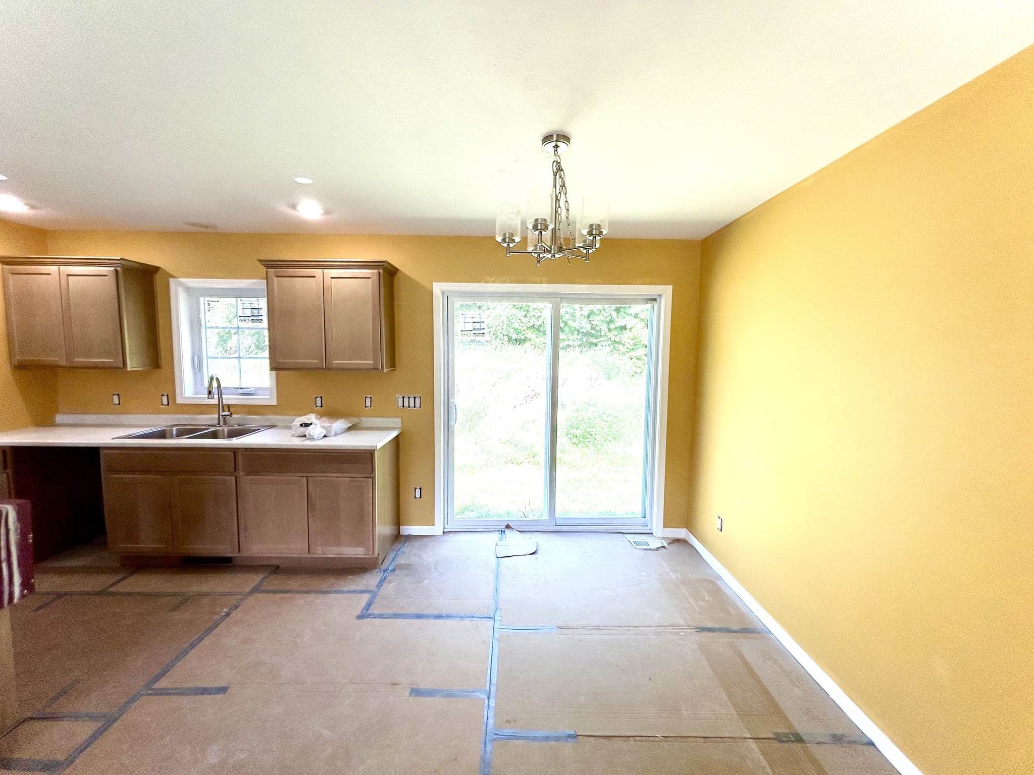 Kitchen interior with warm yellow walls and wooden cabinets. A chandelier hangs over a dining area with sliding glass doors leading outside. Bright and inviting atmosphere.