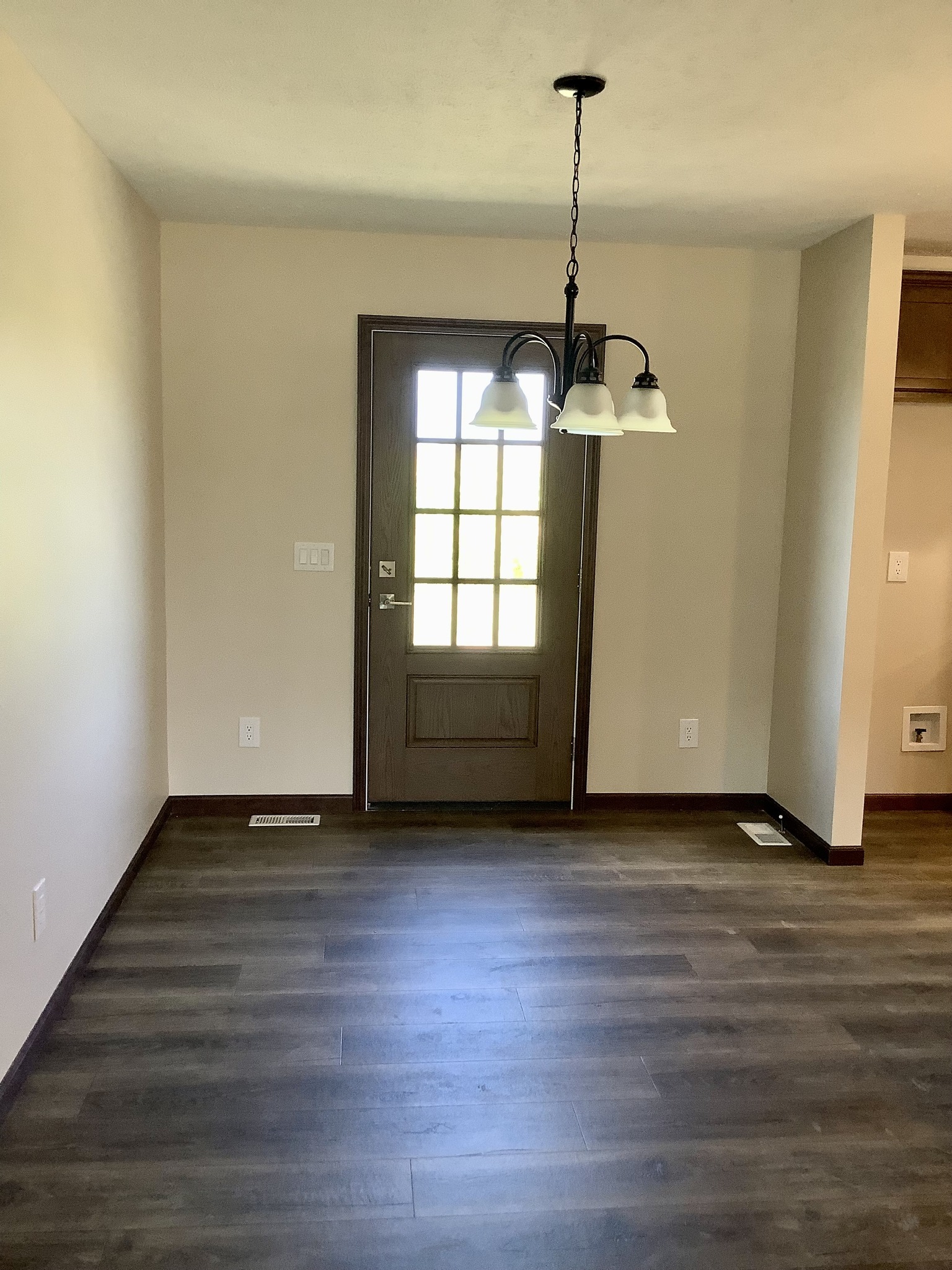 Empty room with a brown wood floor and beige walls. A dark wooden door with glass panels is centered, and a black three-light chandelier hangs above.