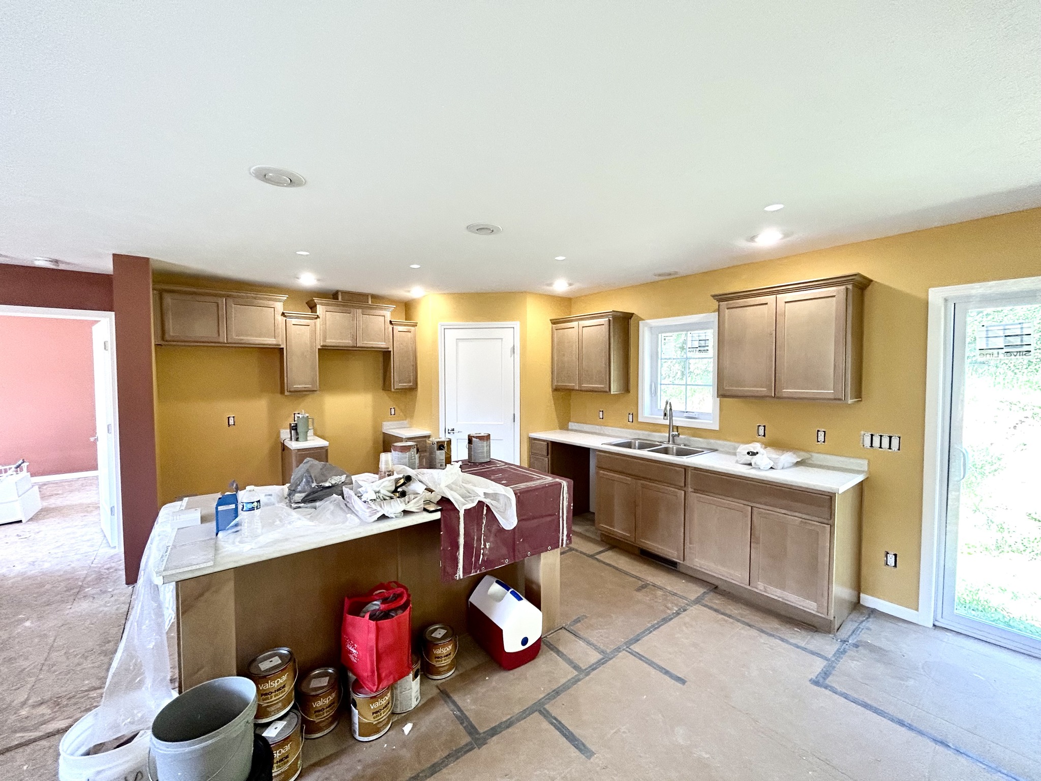 Kitchen under renovation with light wood cabinets, yellow walls, and a central island covered with tools and cloths. Unpainted floor, light spills through windows.