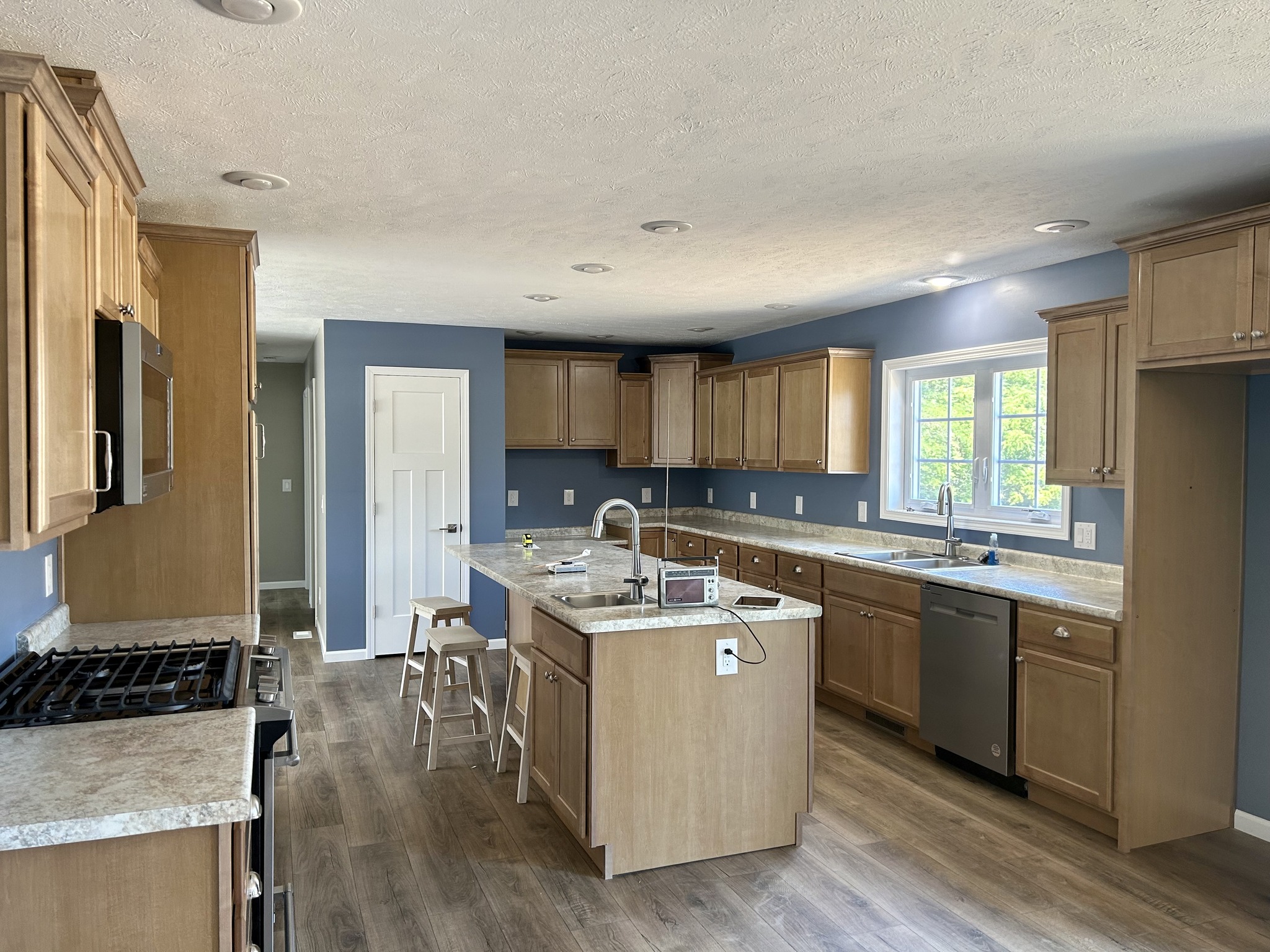 Spacious kitchen with light wood cabinets, marble countertops, and a central island. Blue walls, wood flooring, and bright natural light create a cozy atmosphere.