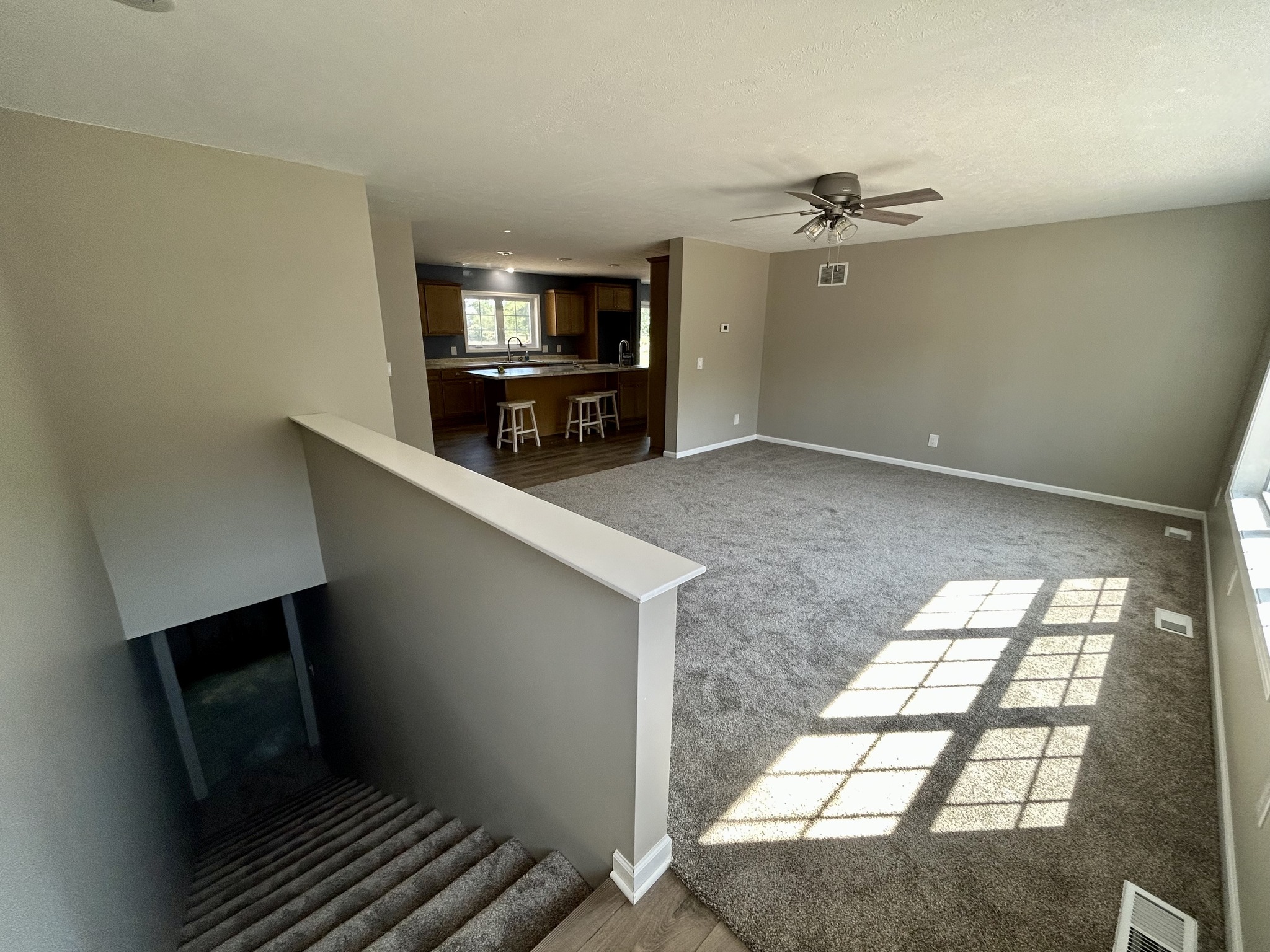 Spacious living area with beige carpet, a ceiling fan, and large windows. Nearby, a wooden kitchen with barstools connects the space. Stairs descend to the left.