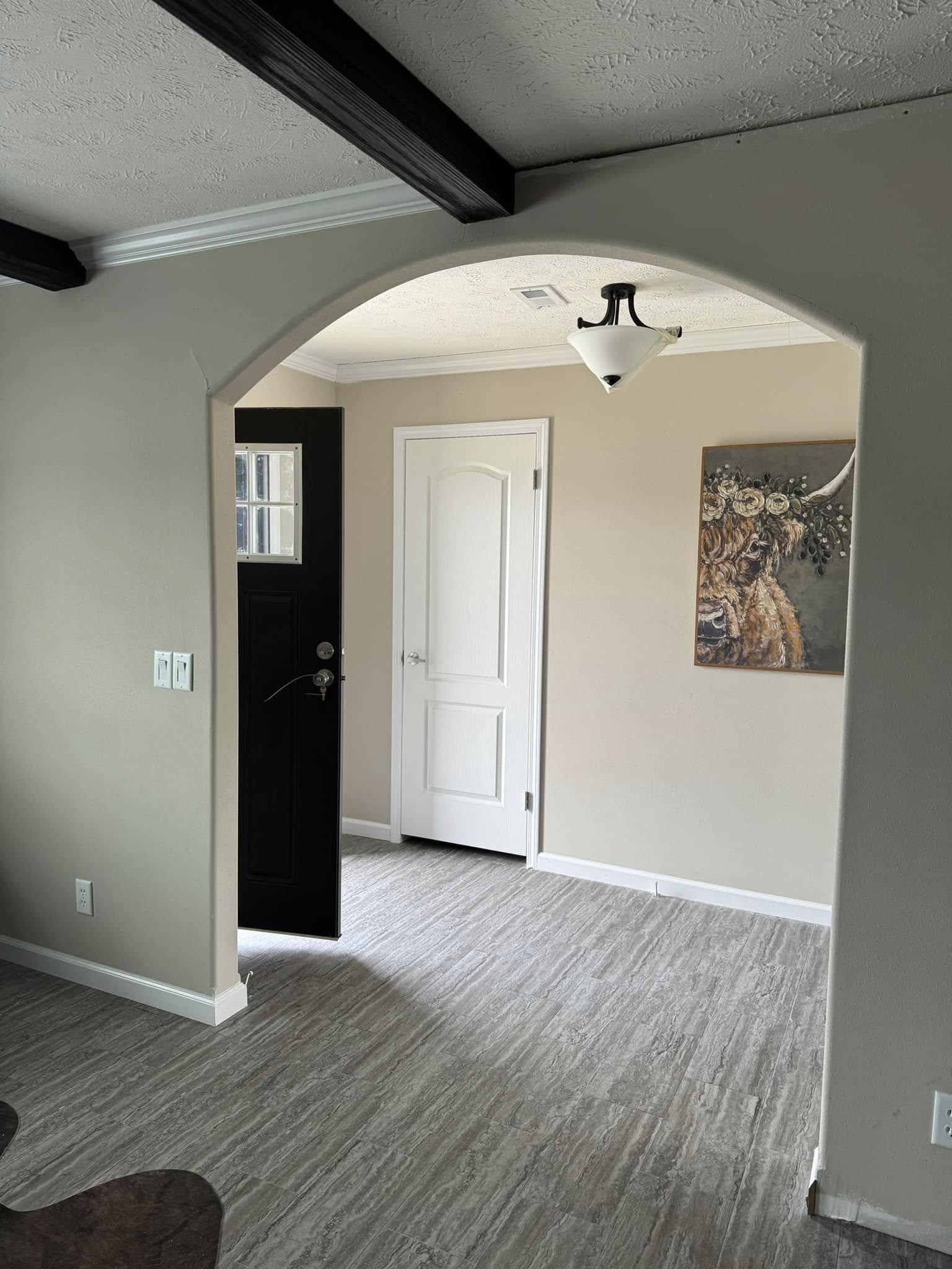A bright, empty room with light wood flooring and beige walls. A black door is ajar, revealing another room featuring a closed white door and a colorful painting of a cow. The ceiling has a decorative black beam and a modern light fixture. The atmosphere is calm and inviting.