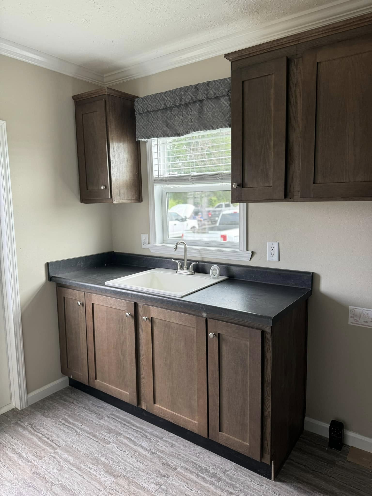 A kitchen features dark wood cabinets with silver knobs above and below a white sink and chrome faucet. A window with a patterned valance is centered above, adding natural light. The floor has a light gray wood pattern. The atmosphere is clean and modern.