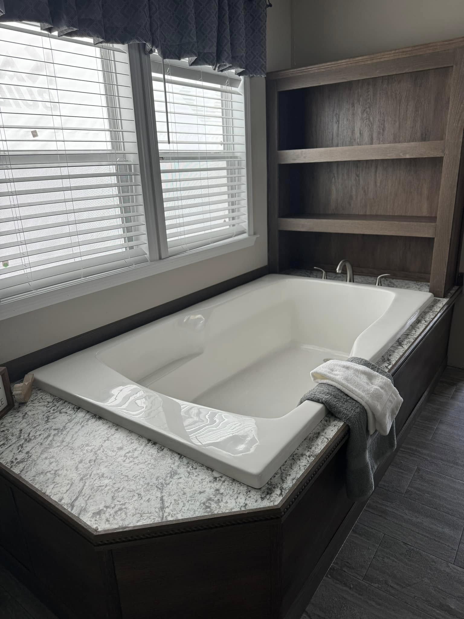 Spacious white bathtub with a marble surround and wood paneling, set beside a large, sunny window with blinds. Towels are neatly draped on the edge.