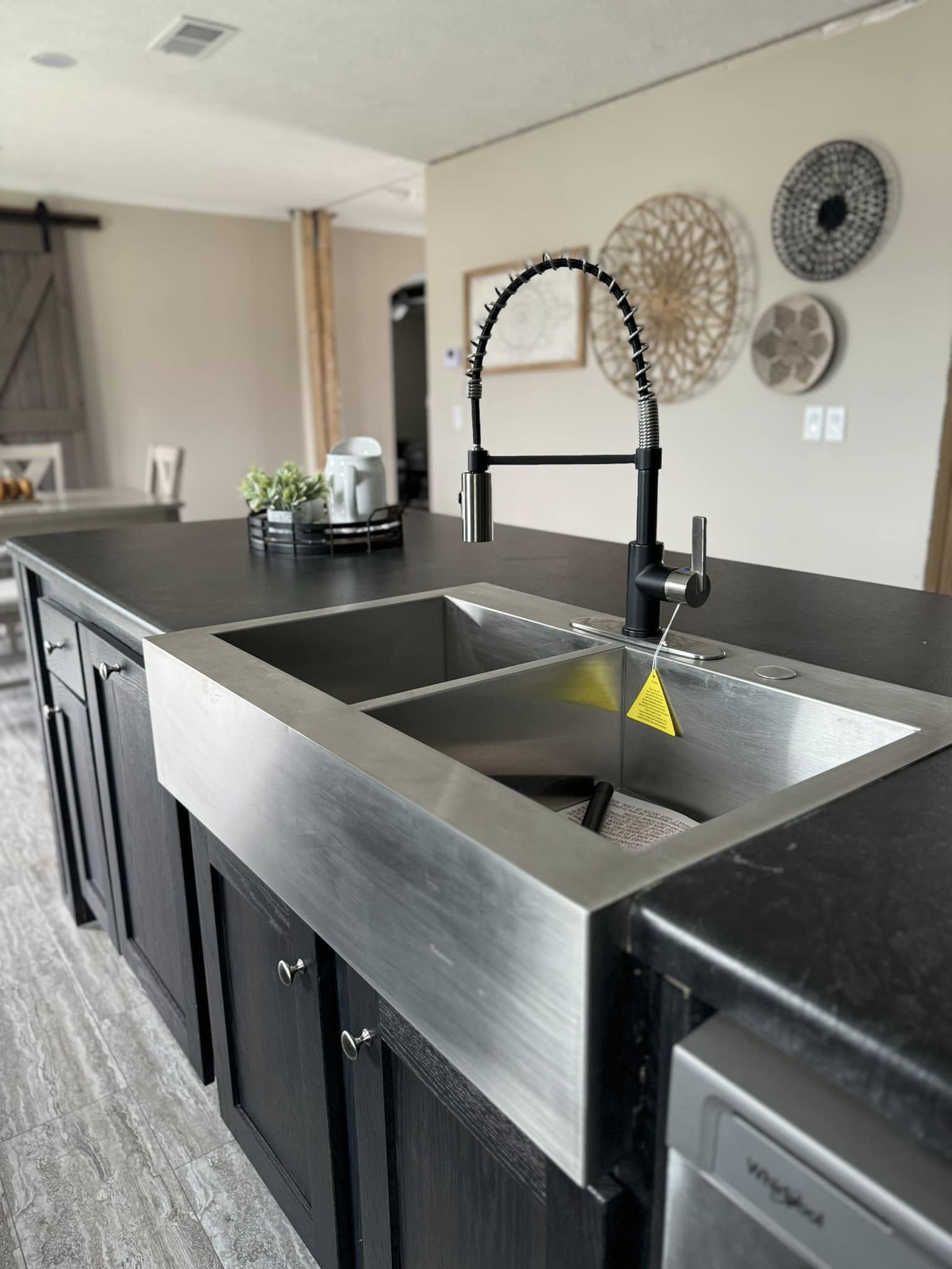 Modern kitchen interior featuring a large stainless steel farmhouse sink with a tall, industrial-style black faucet. The sleek black countertop contrasts with light gray flooring. In the background, wall decor includes woven patterns. The scene conveys a clean, contemporary vibe.