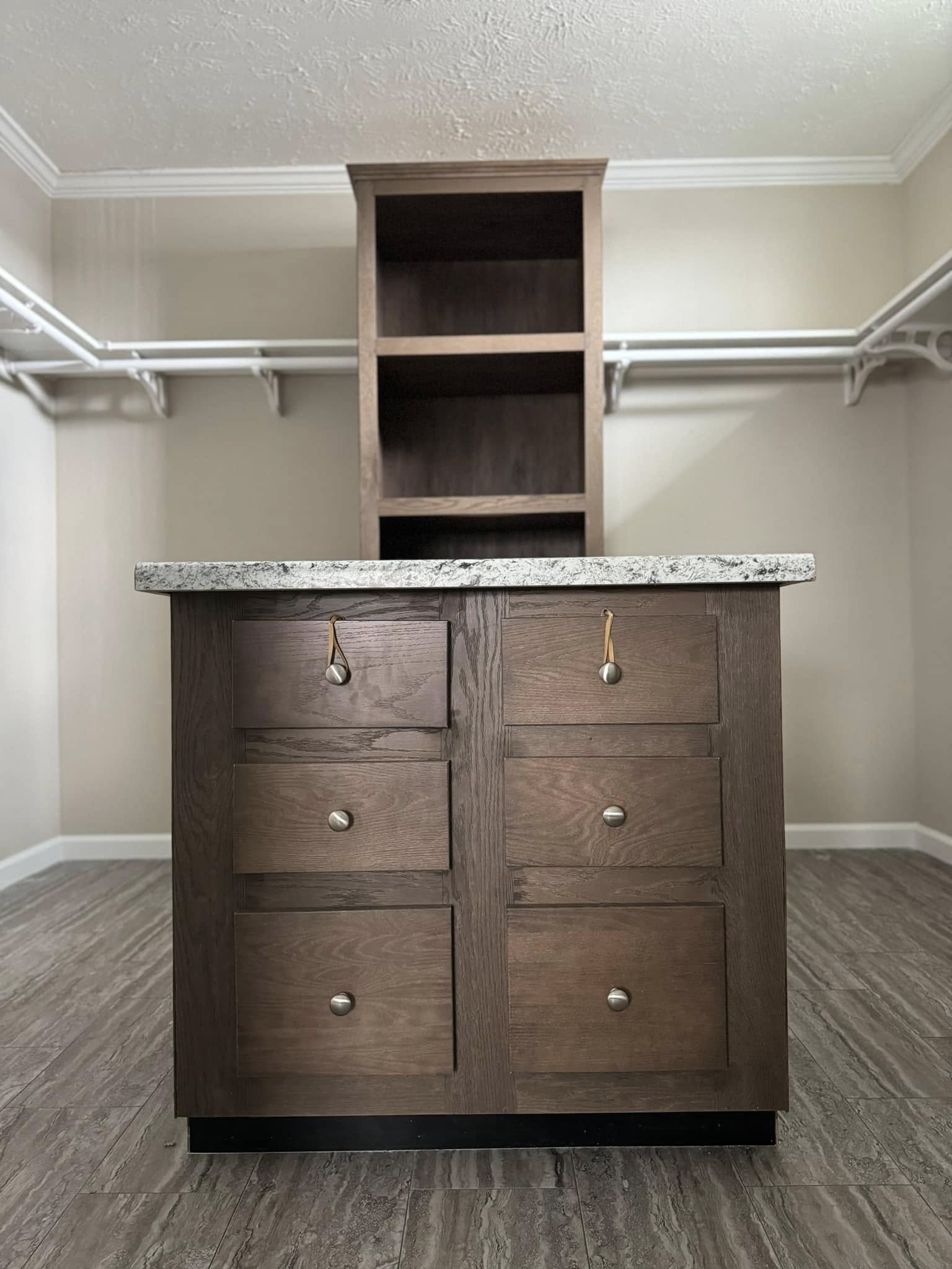 Wooden storage island with six drawers and a speckled granite countertop in a closet with beige walls, shelving, and a modern, organized feel.