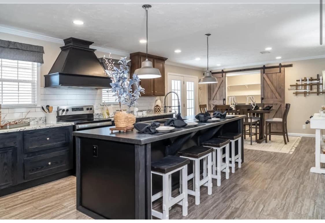 Modern kitchen with a large black island, white stools, and pendant lighting. A dining table is visible in the background near sliding barn doors. Cozy and inviting atmosphere.
