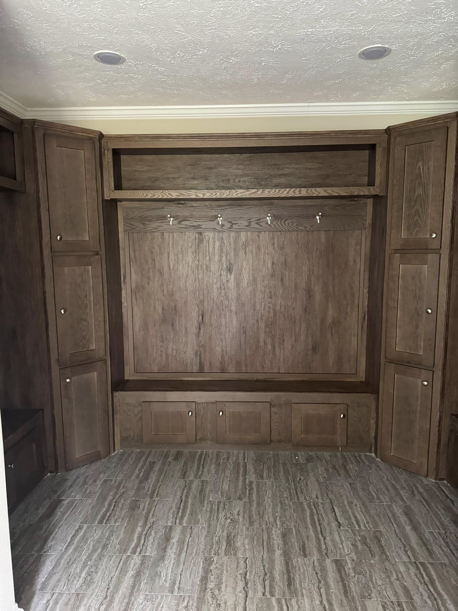 Wooden built-in shelving unit with hooks, cabinets, and storage bench. Surrounding cabinets frame the unit, set against a beige tiled floor.
