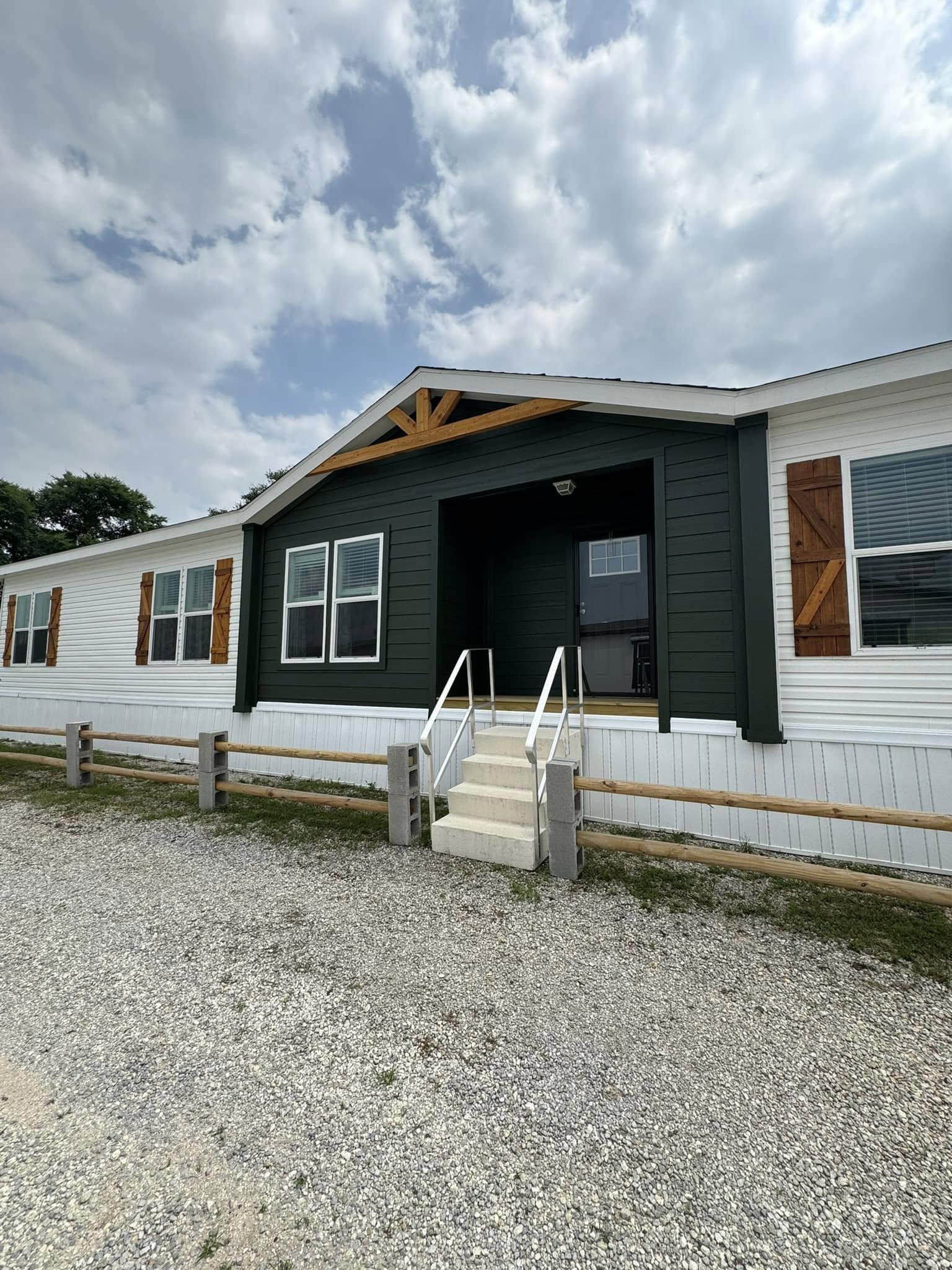 A modern modular home with a dark green facade and white sidings, wooden shutters, and decorative trim. A cloudy sky and gravel path enhance a rustic feel.