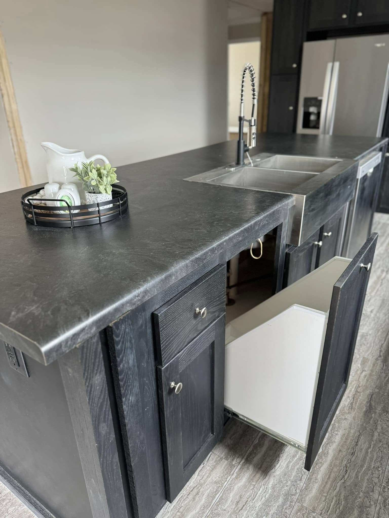 Modern kitchen island with dark countertop and open cabinets. Features a silver sink, tall faucet, and decor tray with a plant, pitcher, and mugs.