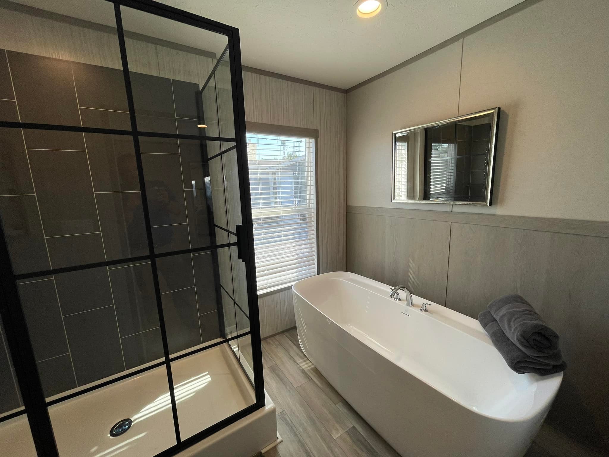 Modern bathroom featuring a white freestanding tub with folded towels, a black-framed glass shower, wood-textured flooring, and a window with blinds.