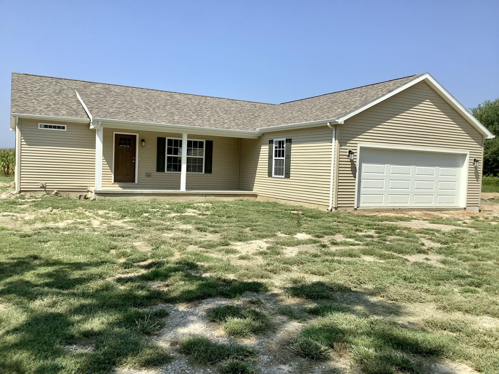 A beige ranch-style house with a covered porch, three windows, and a white garage sits on a patchy lawn under a clear blue sky, conveying a calm, rural feel.