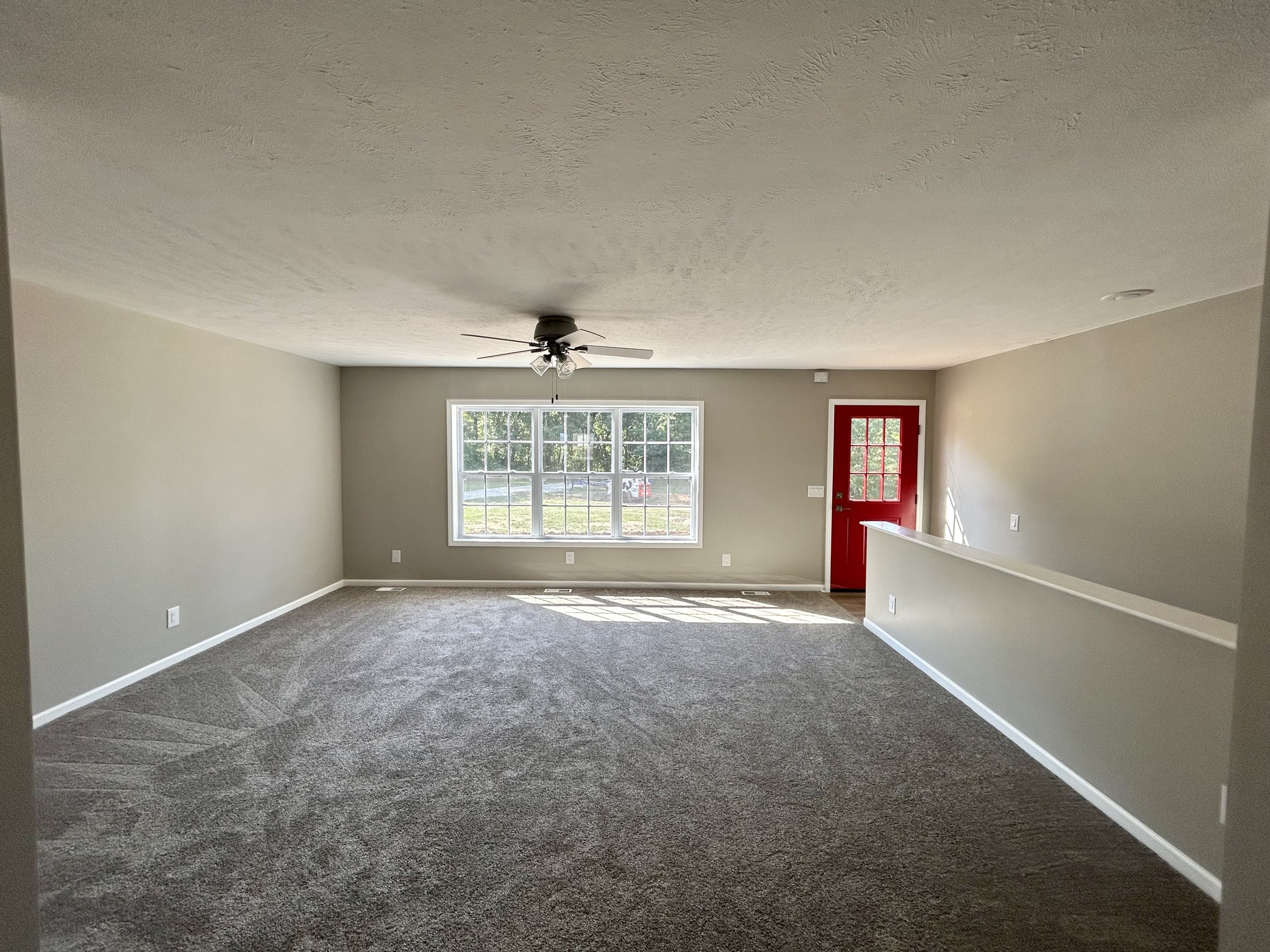 Spacious living room with gray carpet, beige walls, large front window, ceiling fan, and a bold red door, creating a welcoming and airy atmosphere.
