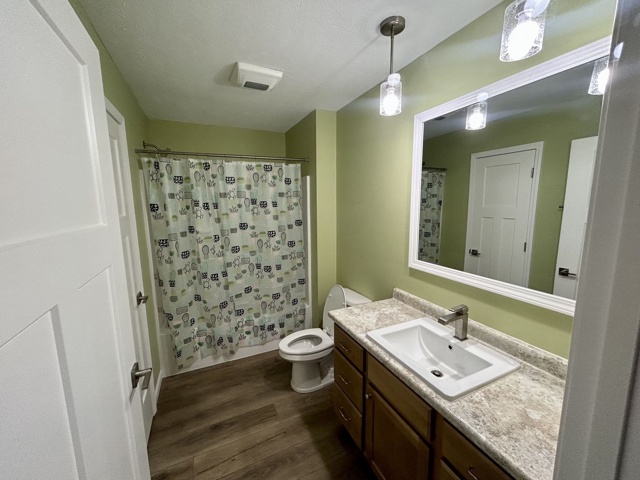 Modern bathroom with green walls and wood floors. It features a white sink with a spacious countertop, a patterned shower curtain, and bright lighting.