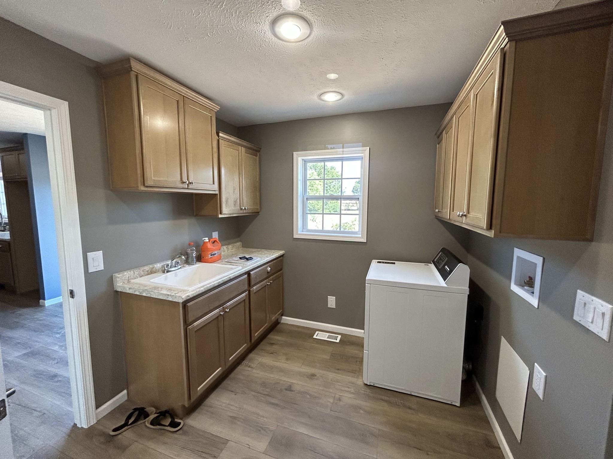 A small laundry room with wooden cabinets, a white washer, and a deep sink. Soft lighting and a window create a cozy, functional atmosphere.