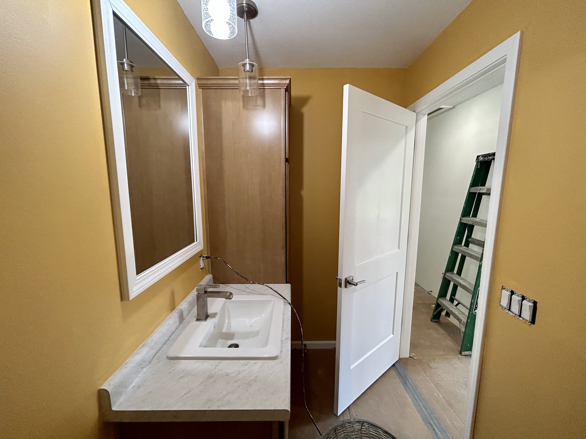 Small bathroom with mustard-yellow walls, a white sink, and wooden cabinets. Open door reveals a green ladder in the adjacent room, suggesting ongoing renovation.