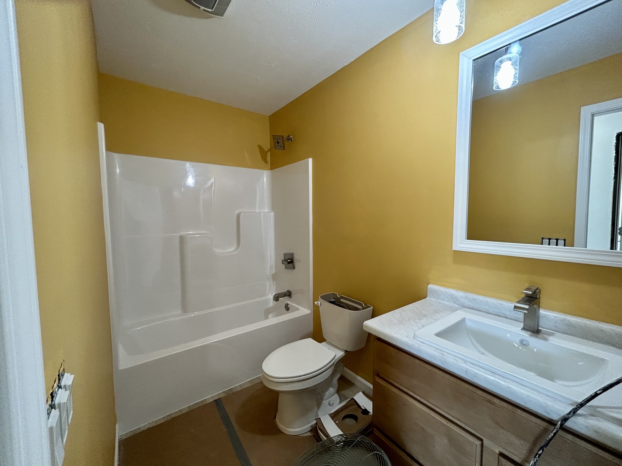 A small bathroom with yellow walls features a white bathtub, a toilet, and a vanity with a marble sink. A large mirror reflects light, creating a warm and inviting atmosphere.