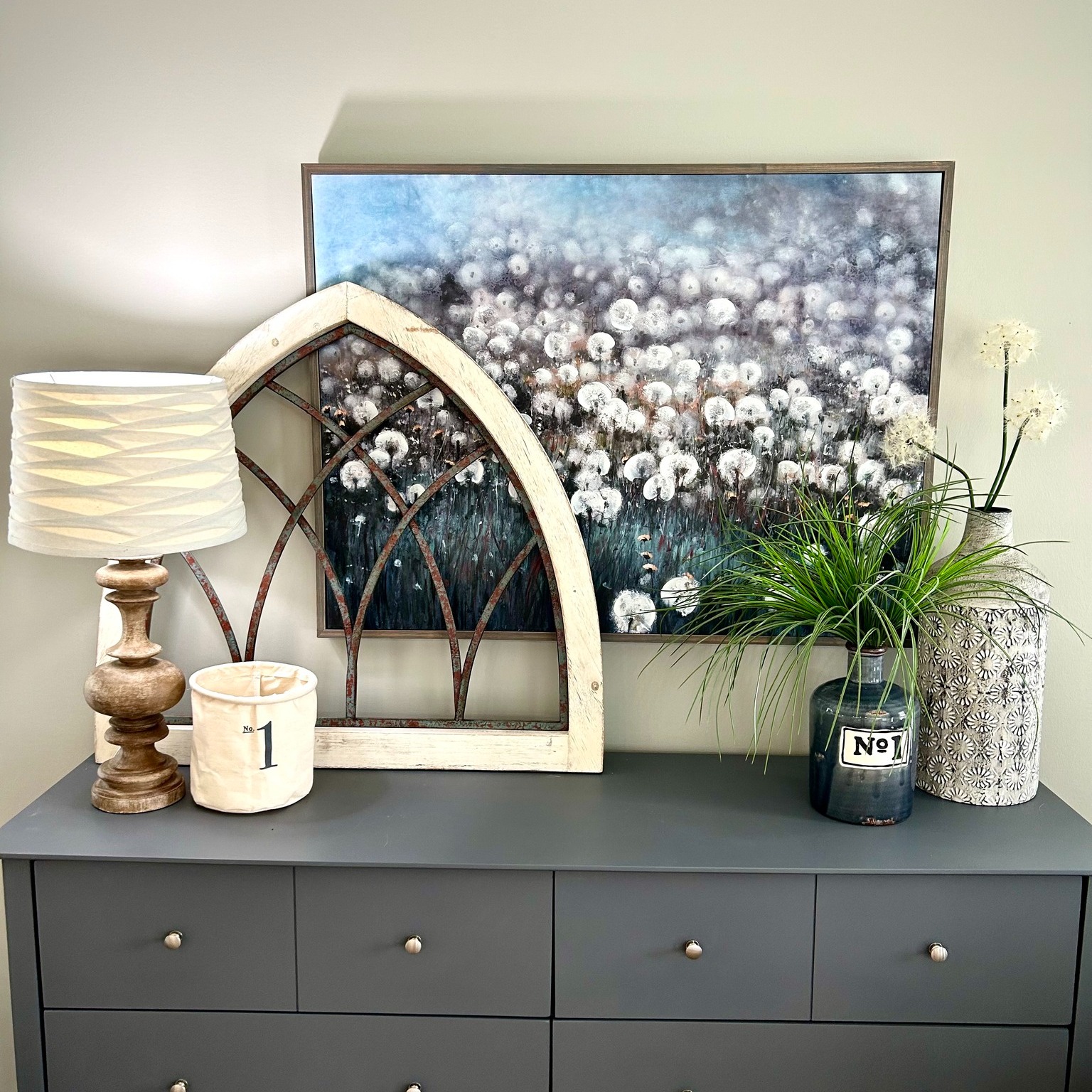 A decorative arrangement on a gray dresser features a modern lamp, a white frame, dandelion artwork, and vases of greenery, creating an elegant, serene atmosphere.