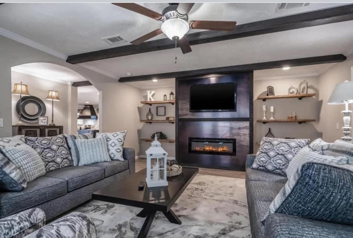 Modern living room with grey sofas, patterned cushions, and a black coffee table centered by a white lantern. A wall-mounted TV and fireplace, flanked by decorative shelves, create a cozy atmosphere. Ceiling fan and elegant lighting enhance the warm, inviting tone.