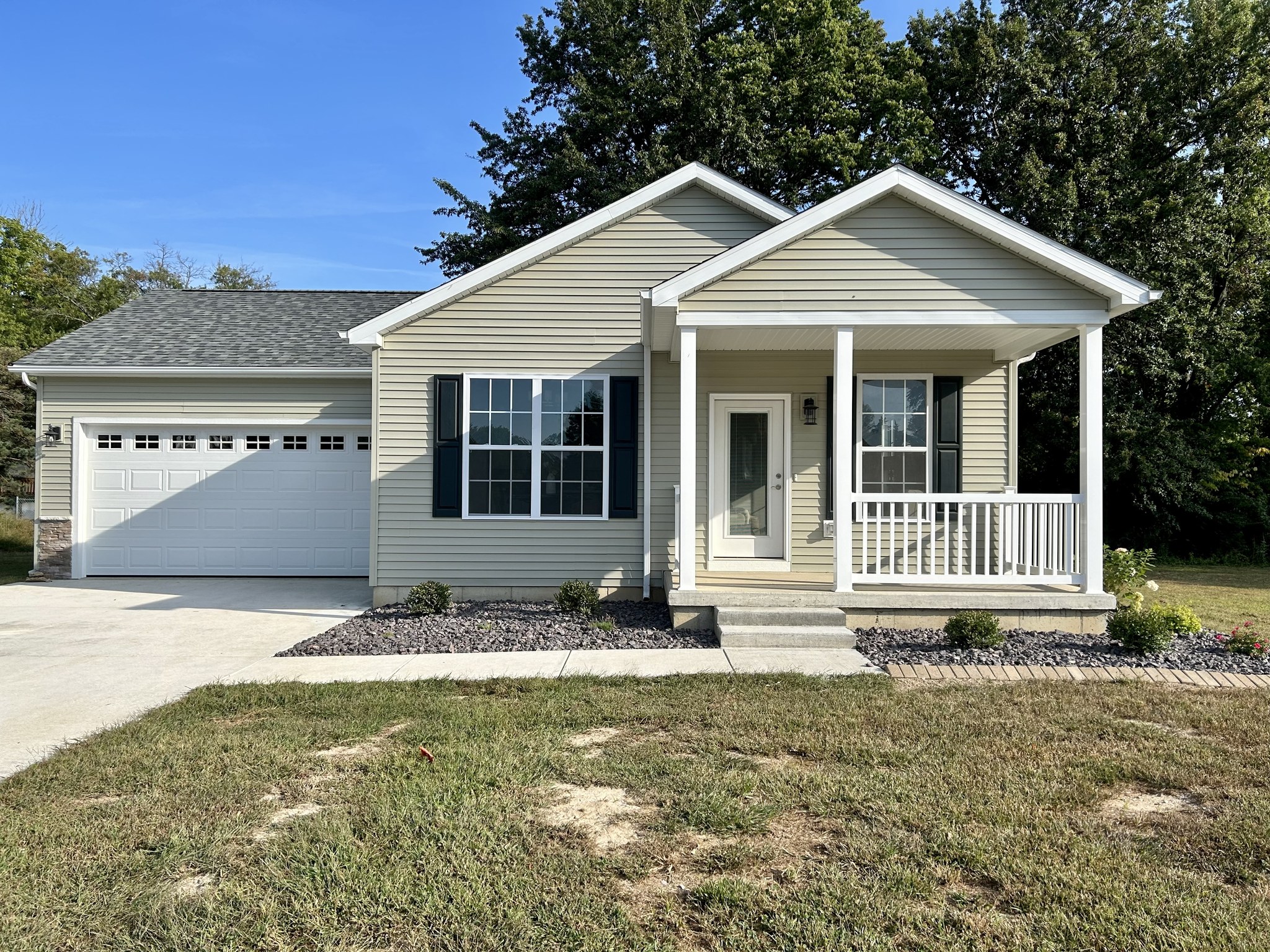 A cozy single-story house with beige siding, a covered porch, and two white garage doors. It's surrounded by greenery under a clear blue sky.