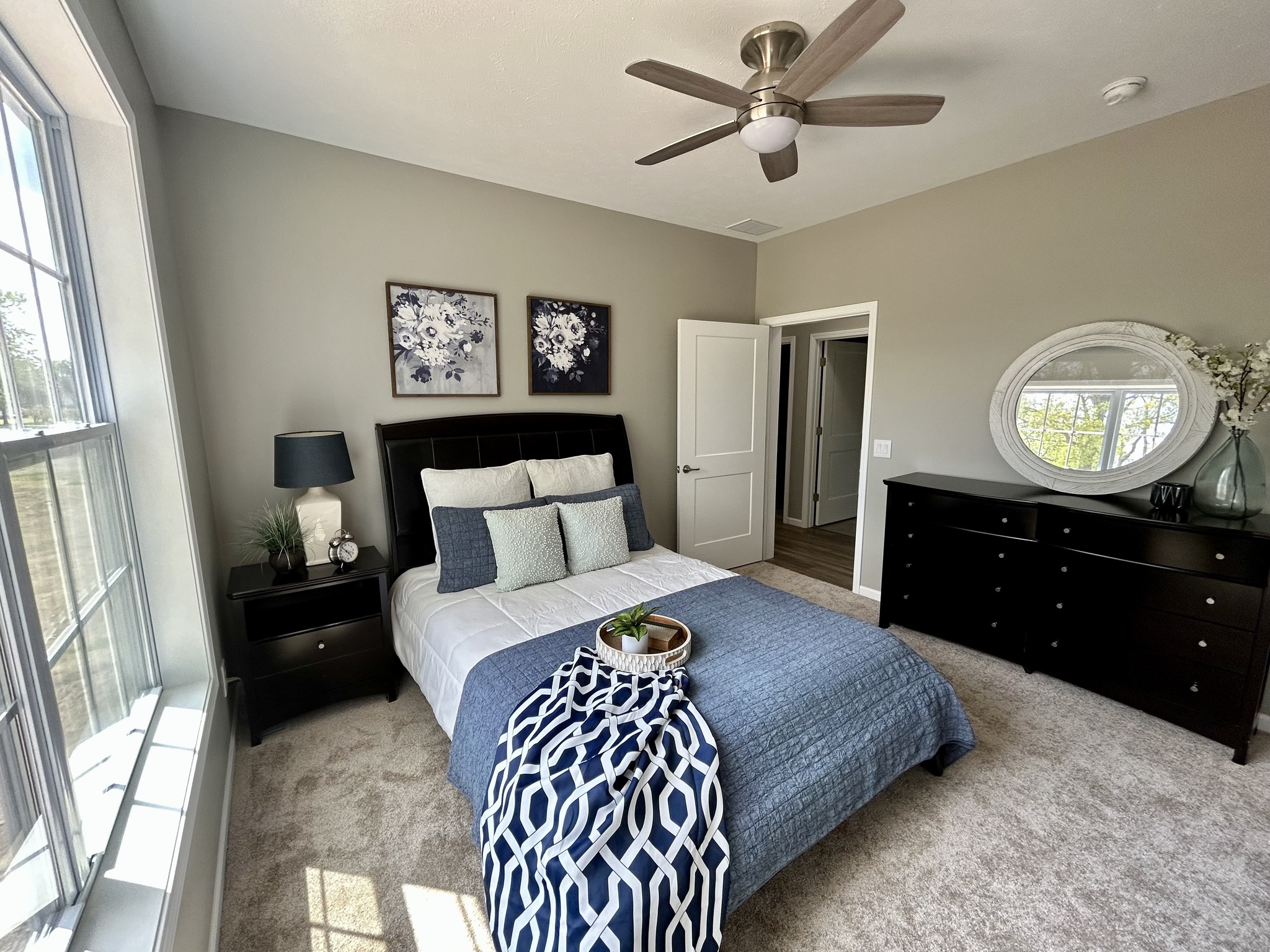 Bright bedroom with large window and ceiling fan. Features a bed with blue and white linens, two floral paintings, and a dark dresser with an oval mirror. Cozy and elegant atmosphere.