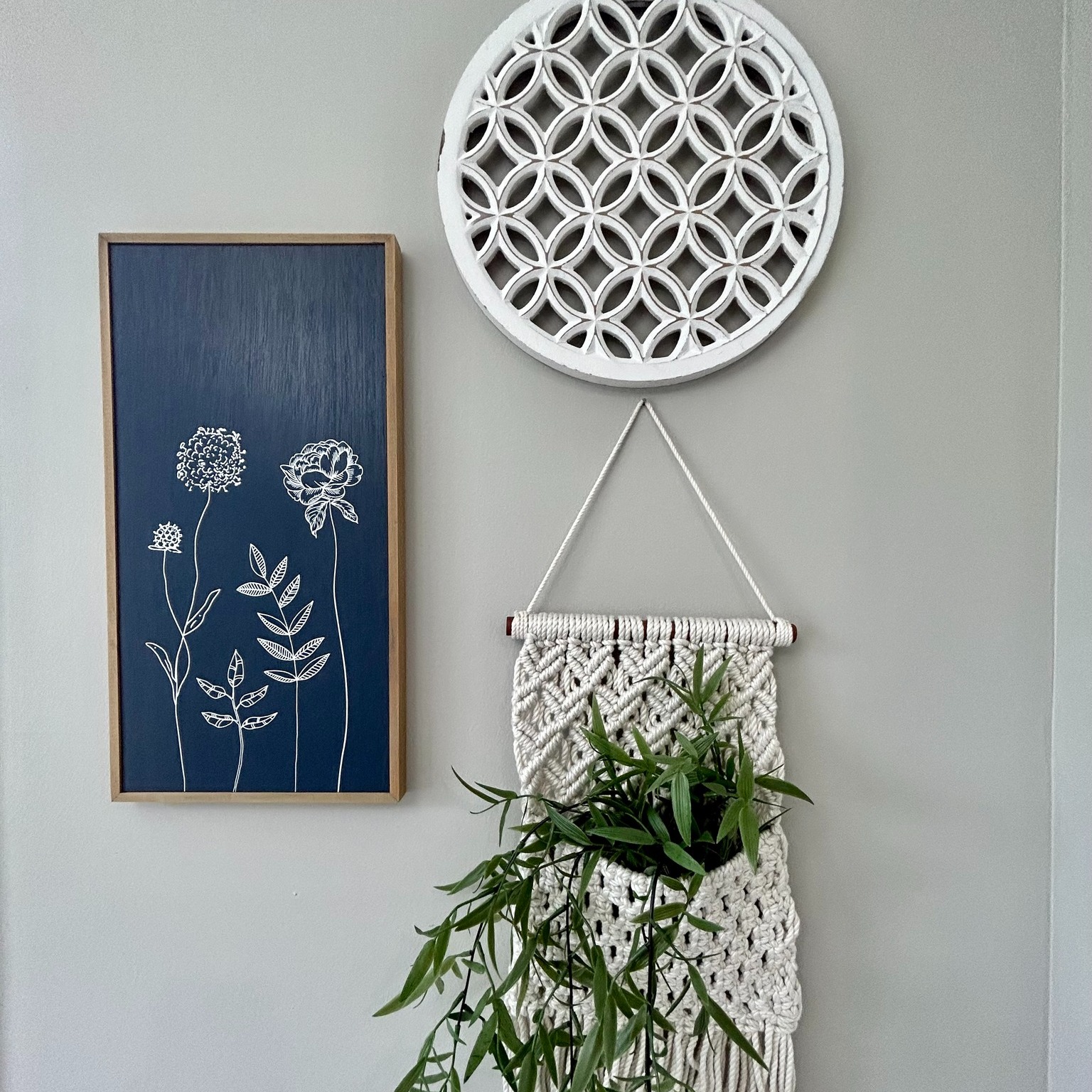Minimalist wall display with a round white lattice, a blue floral line art, and a macramé planter with green leaves, on a light gray background.