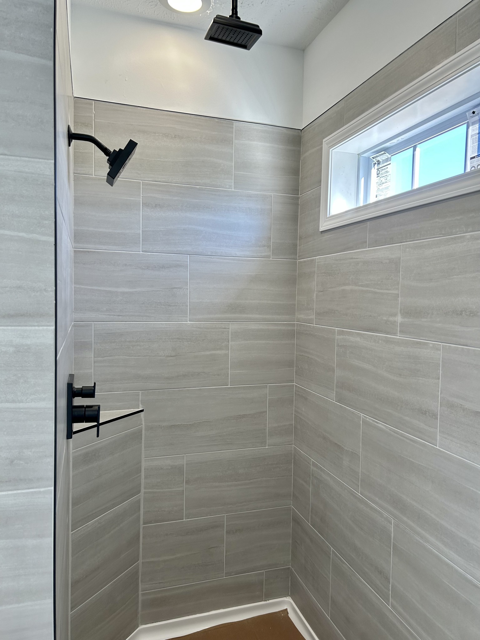 Minimalist shower with large beige tiles, a black rain showerhead, and a small rectangular window. The bright, clean setting feels modern and serene.