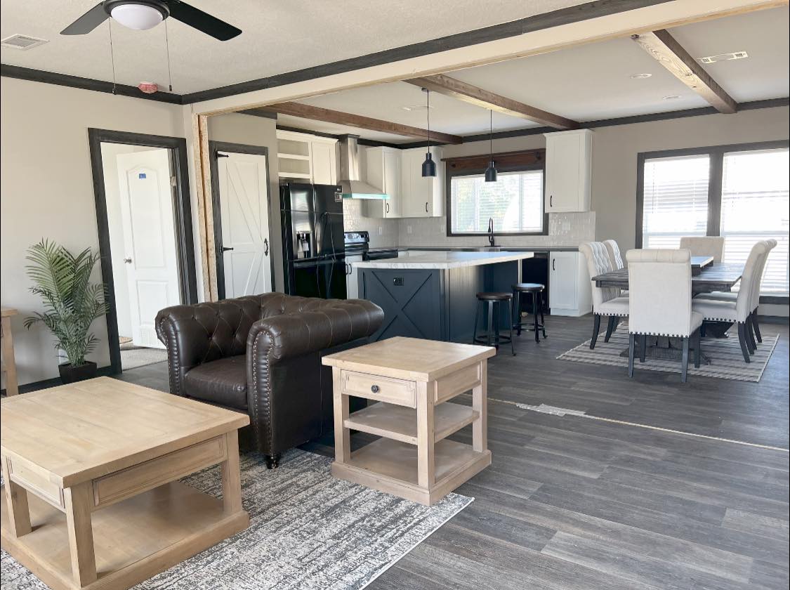Modern open-concept living area with a brown leather armchair, light wood tables, kitchen island with black appliances, and dining table. Bright and airy atmosphere.