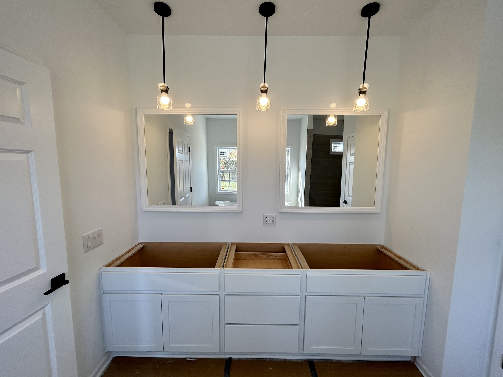 Modern bathroom under construction features two framed mirrors above a double sink vanity area with no sinks. Three pendant lights illuminate the space.