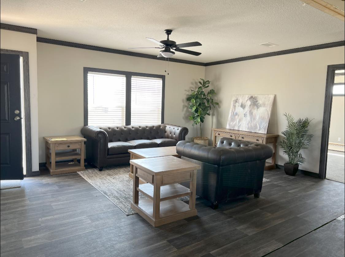 A modern living room with two dark leather sofas, a wooden coffee table, and side tables. Large windows, potted plants, and a ceiling fan enhance the cozy ambiance.