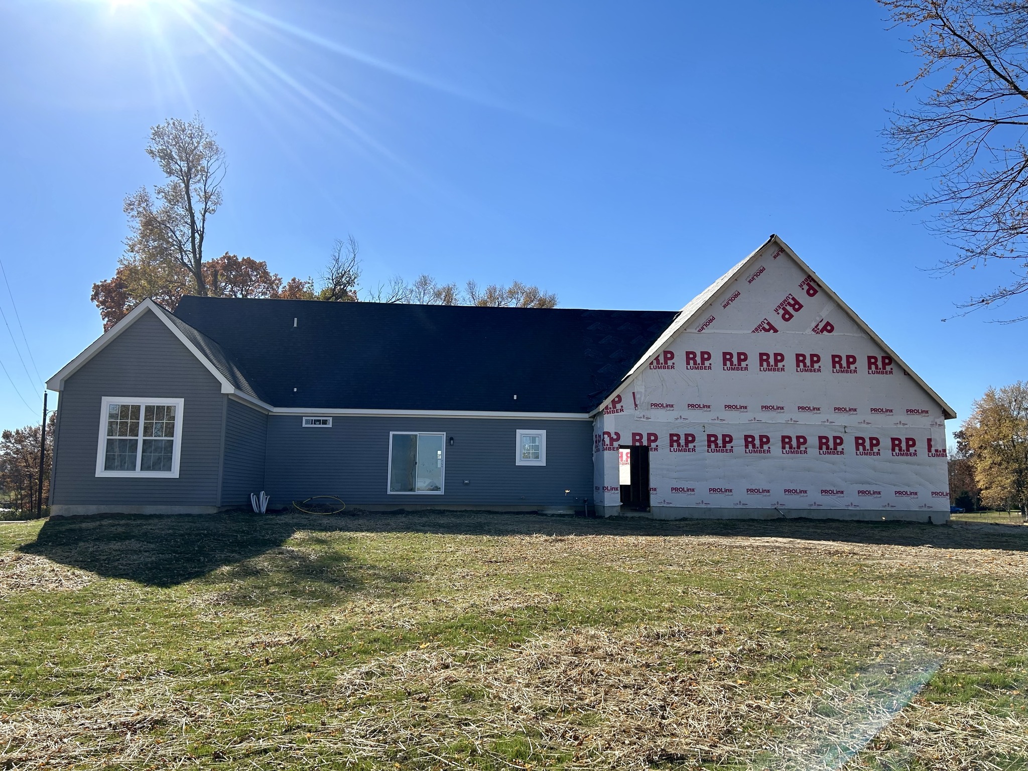Single-story house under construction, with gray siding, unfinished areas covered in Tyvek wrap. Sunlit, grassy yard, and autumn trees in the background.