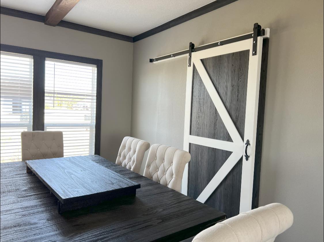 Dining room with a rustic tone, featuring a wooden table, cream upholstered chairs, a large sliding barn door with a white frame, and a sunlit window.