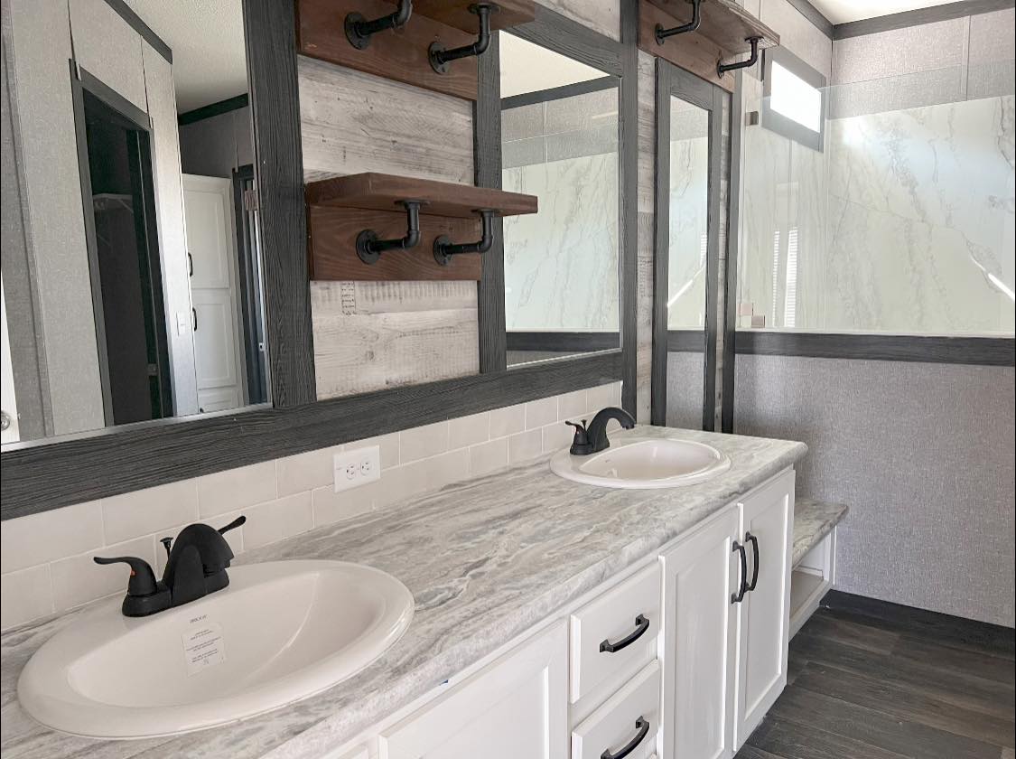 Modern bathroom with dual sinks on a marble countertop. White cabinets, dark faucets, large mirror, and rustic wooden shelves create a sleek, calming atmosphere.