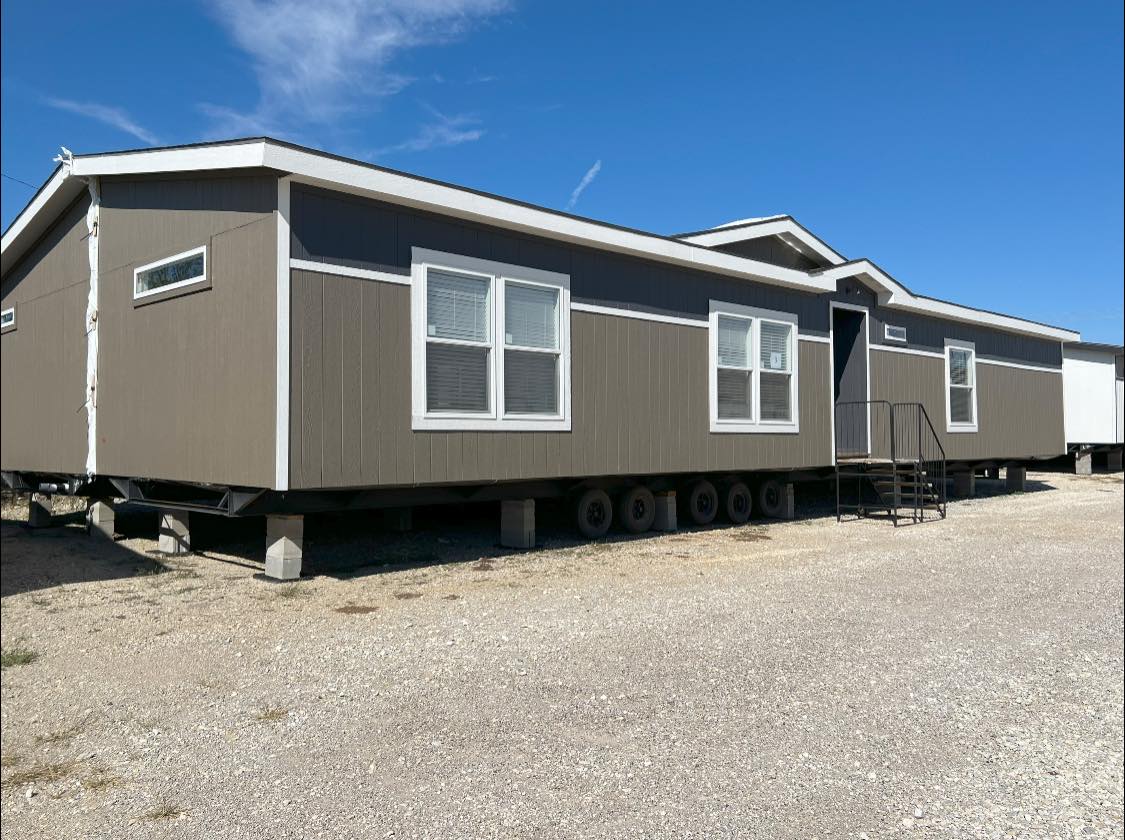 Brown manufactured home on a gravel lot under a clear blue sky. It has white-trimmed windows, a small staircase, and sits on wheels, ready for transport.