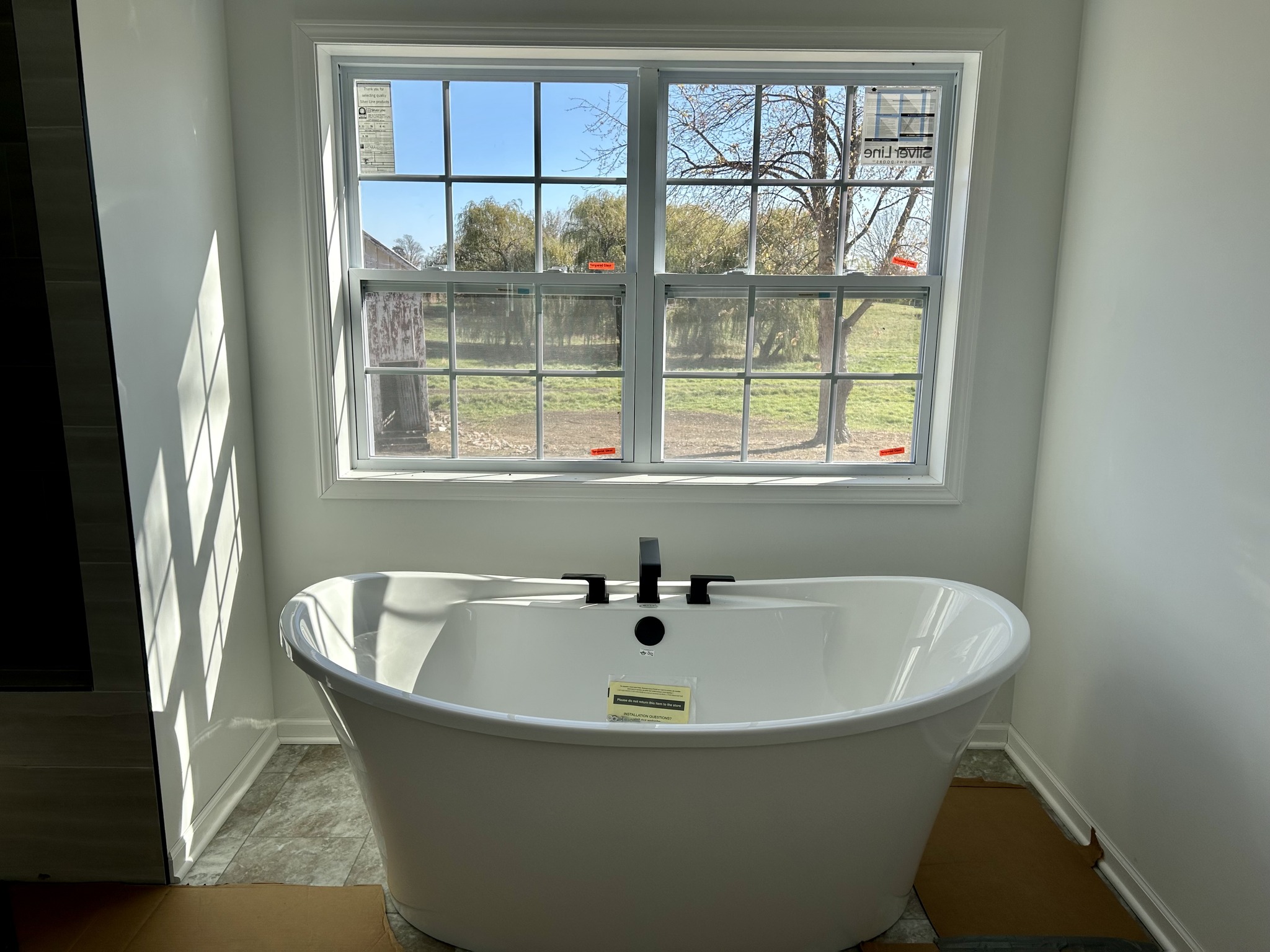 Freestanding white bathtub beneath a large window with a sunny outdoor view. The room features neutral walls, creating a tranquil and inviting atmosphere.