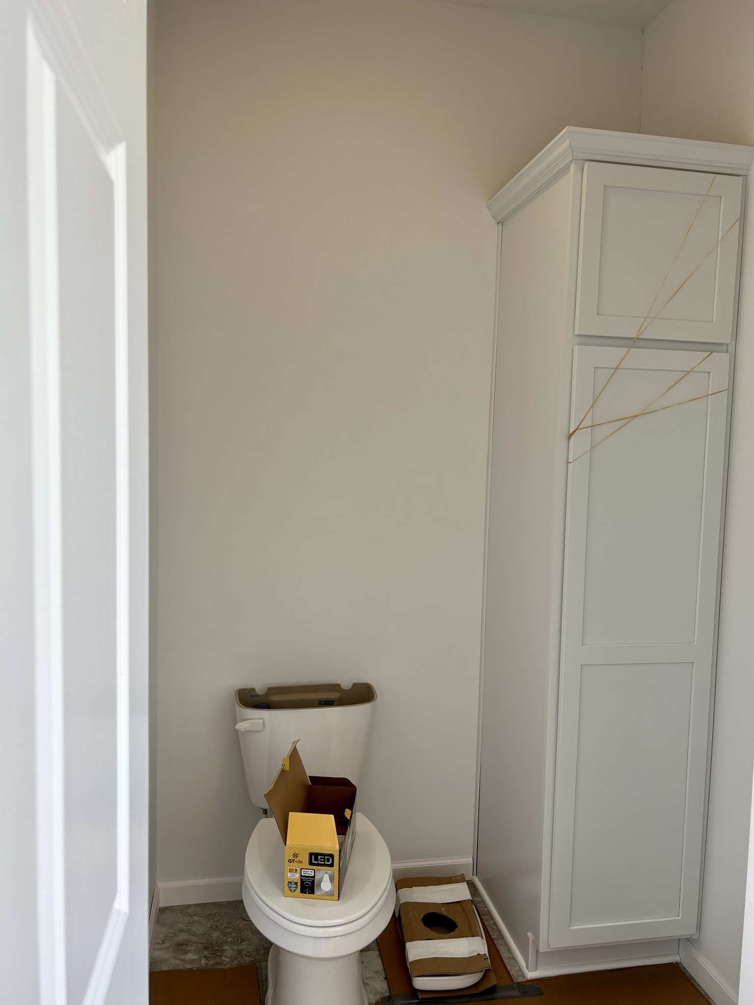 Small bathroom with a toilet, open LED light bulb box on the seat, and tall white cabinet. Minimalist decor, slightly cluttered with packaging.