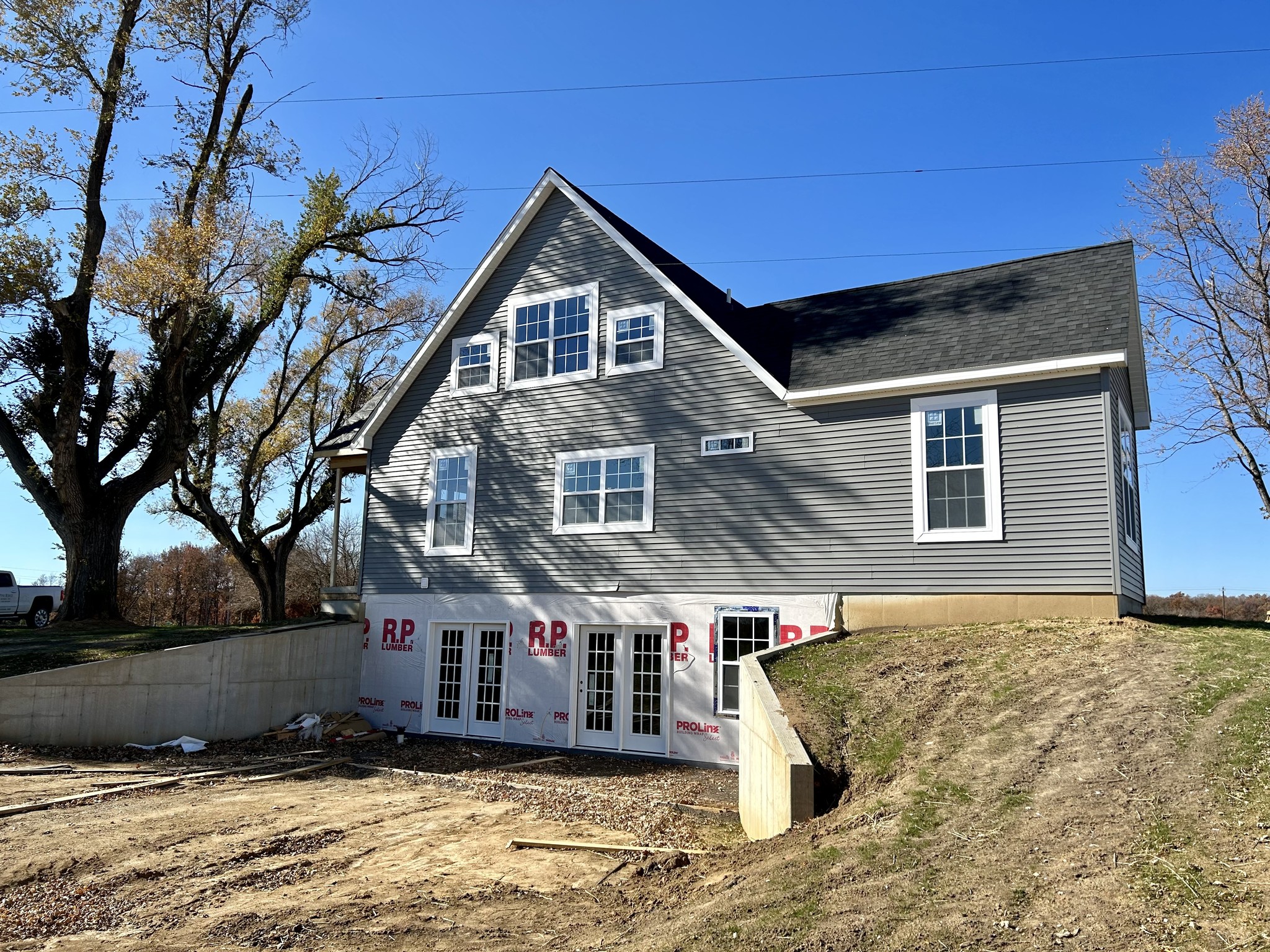 A newly constructed house with gray siding and white trim, featuring large windows and a steep roof. It's set against a clear blue sky, with surrounding bare trees.