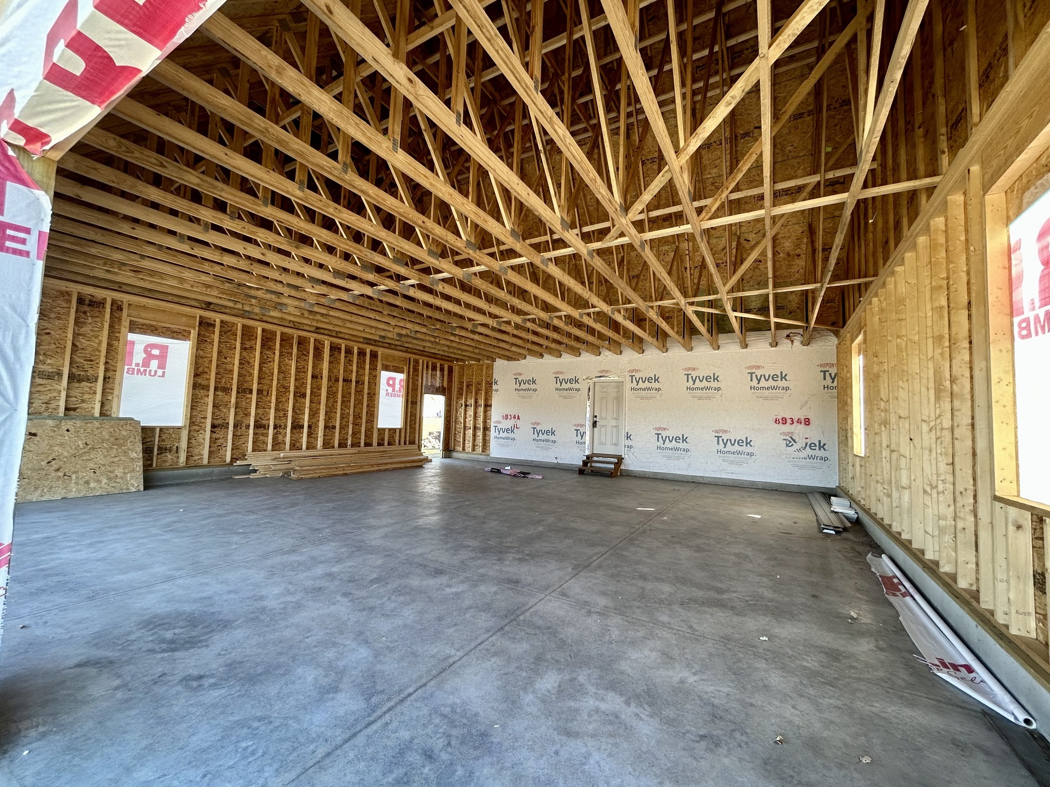 Spacious unfinished building interior with exposed wooden beams and plywood walls. Bright natural light filters through multiple openings, creating an airy atmosphere.
