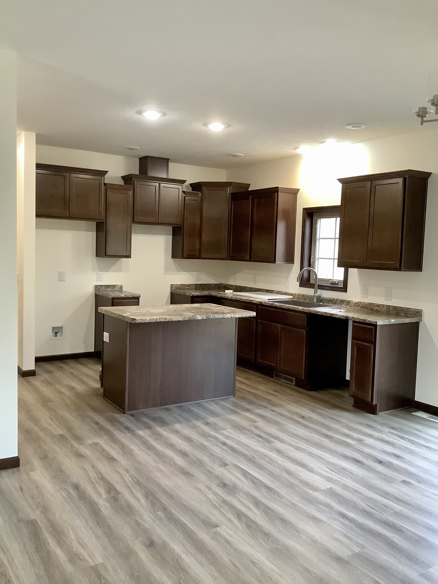 Modern kitchen with dark wooden cabinets, a central island, and marble countertops. The wood-look flooring adds warmth, illuminated by ceiling lights.