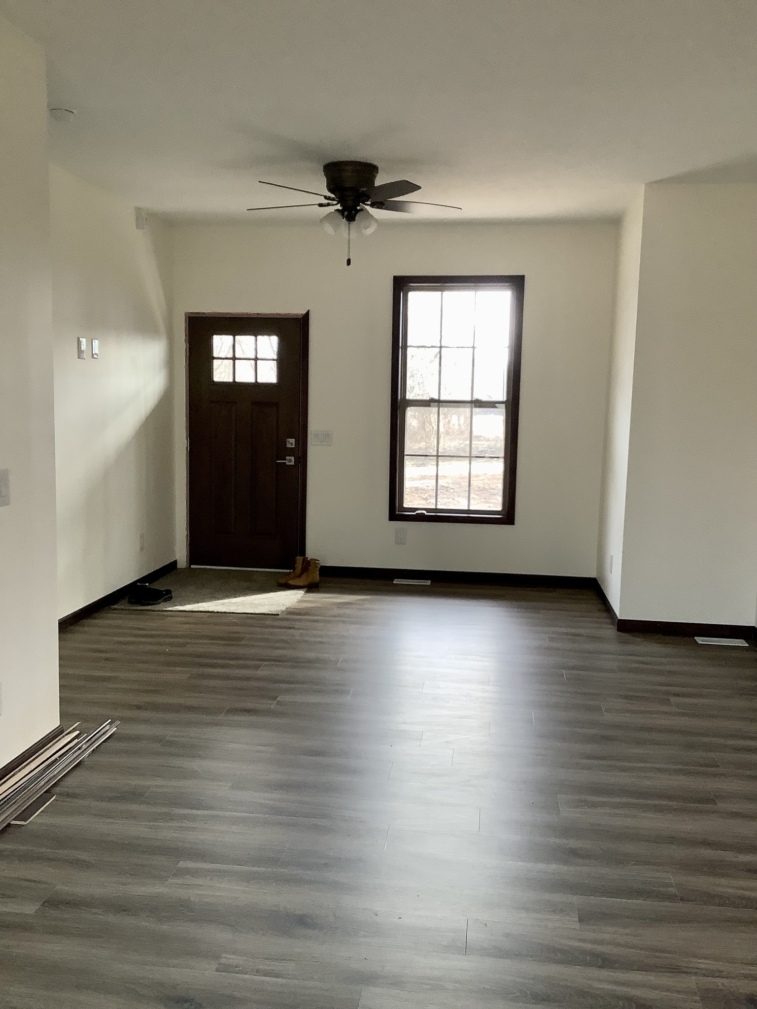 A vacant room with light wood flooring, a dark brown door, and a window letting in natural light. Ceiling fan above; minimalist, calm atmosphere.